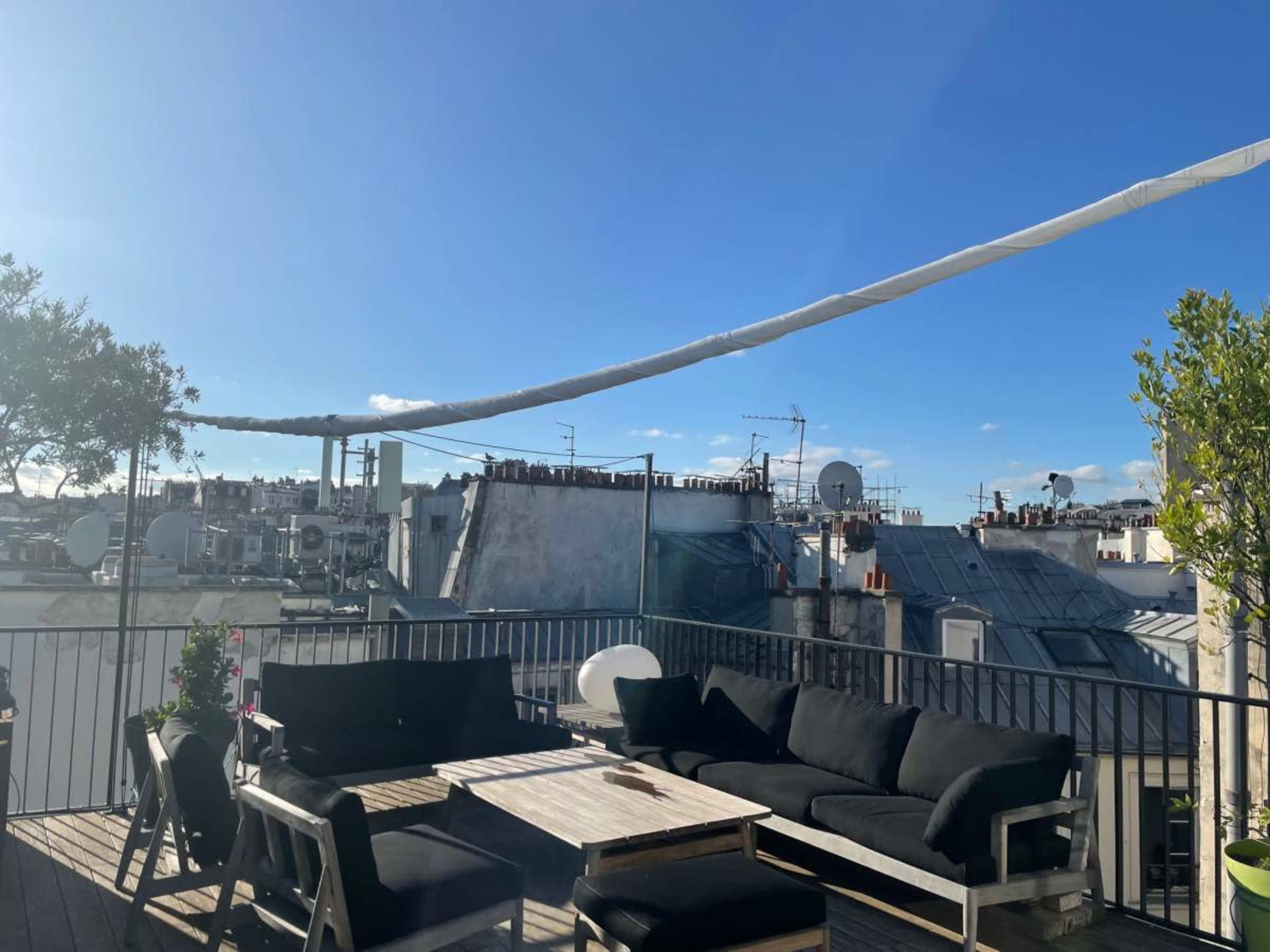 The image shows a rooftop terrace with a black sofa set and a view of rooftops and antennas under a blue sky.