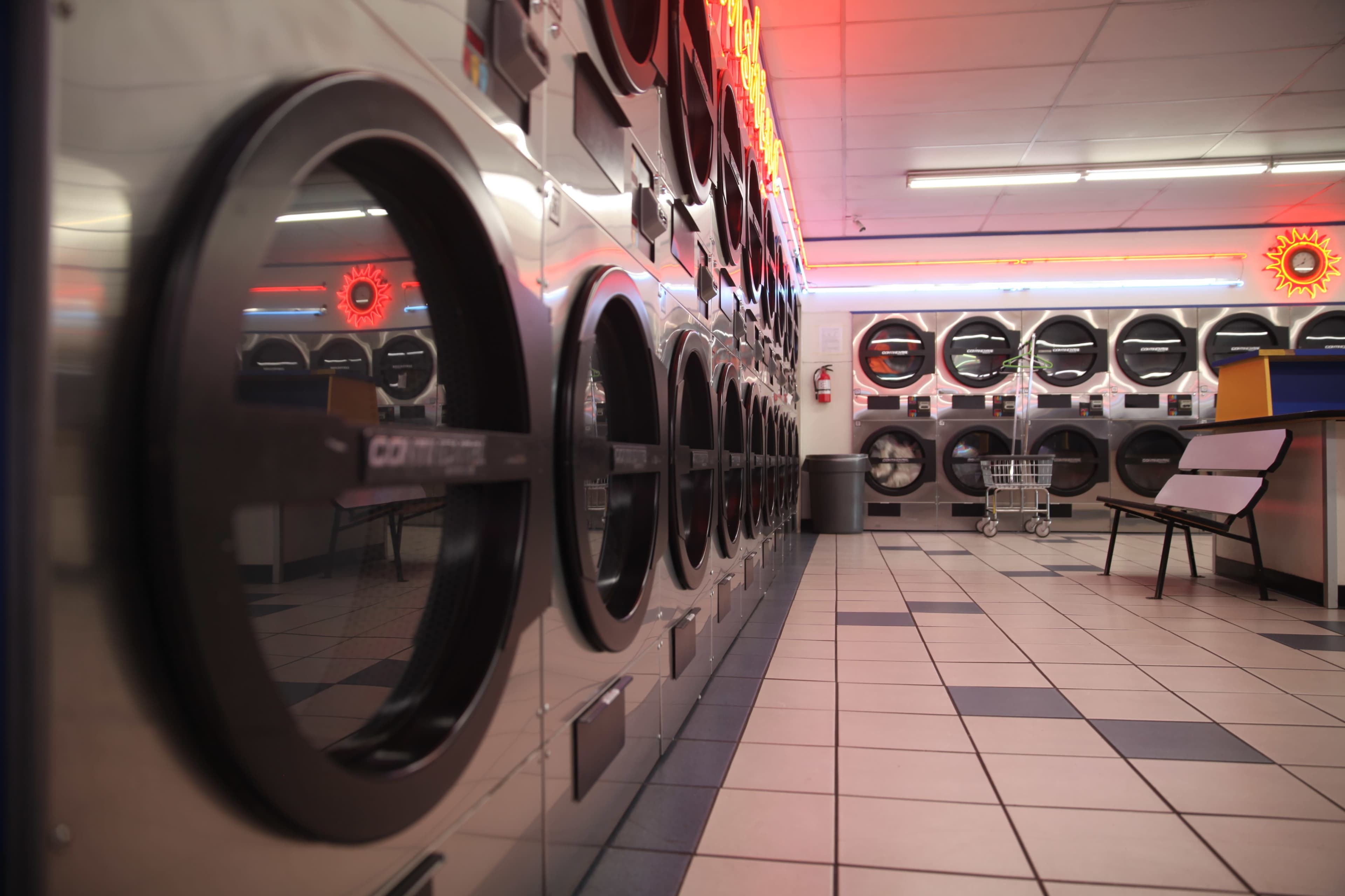 The image shows an interior view of a laundry facility with rows of industrial washing machines and dryers, illuminated by overhead lights.