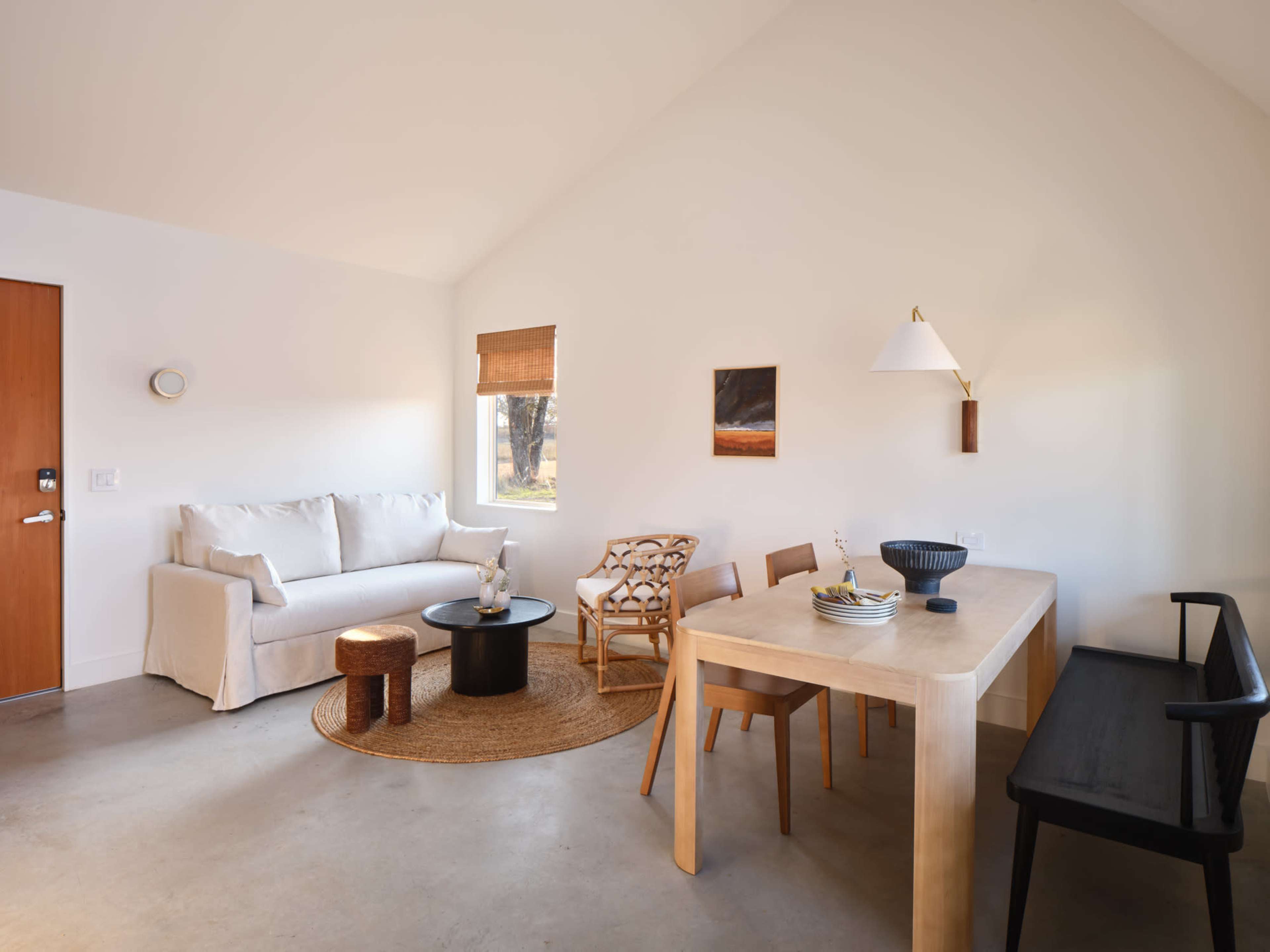 A modern living space features a white sofa, a round black coffee table, a wooden dining table with chairs, and minimal decor against a neutral backdrop.