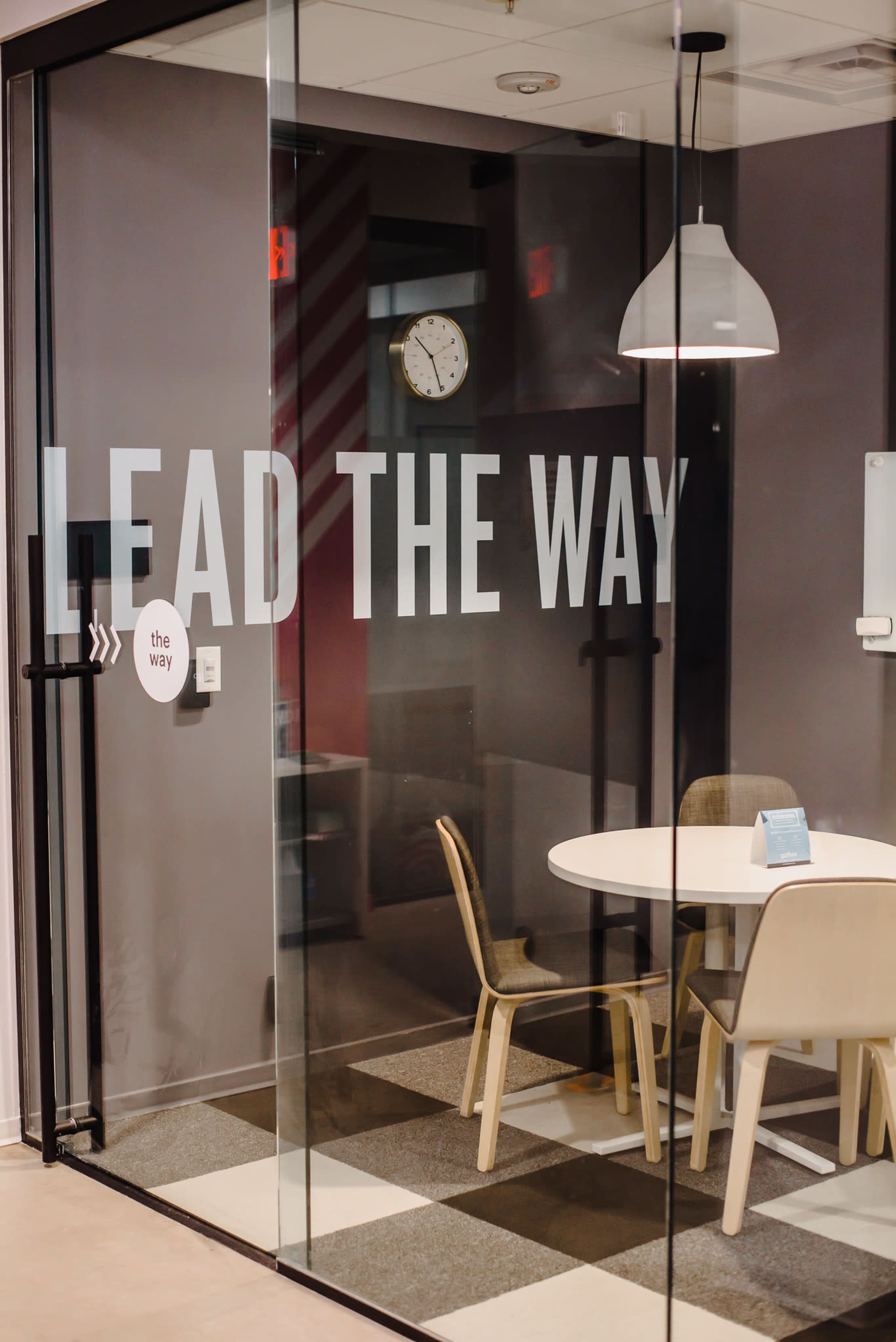 The image shows a glass-walled meeting room with the phrase "LEAD THE WAY" displayed prominently, a round table, and two chairs.