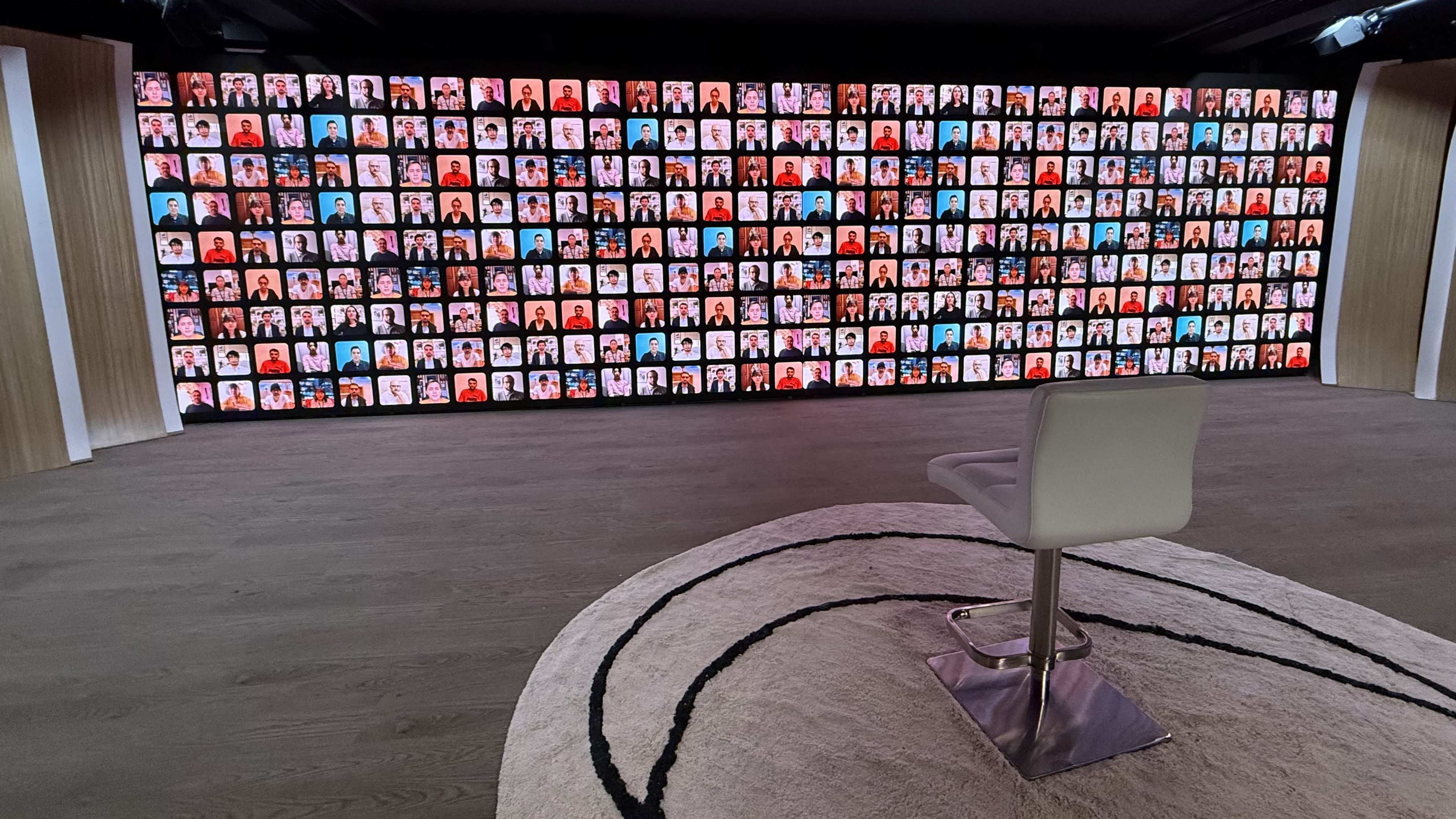 The image shows a large display wall featuring numerous individual portraits, with a single empty chair positioned in front of a circular rug on the floor.