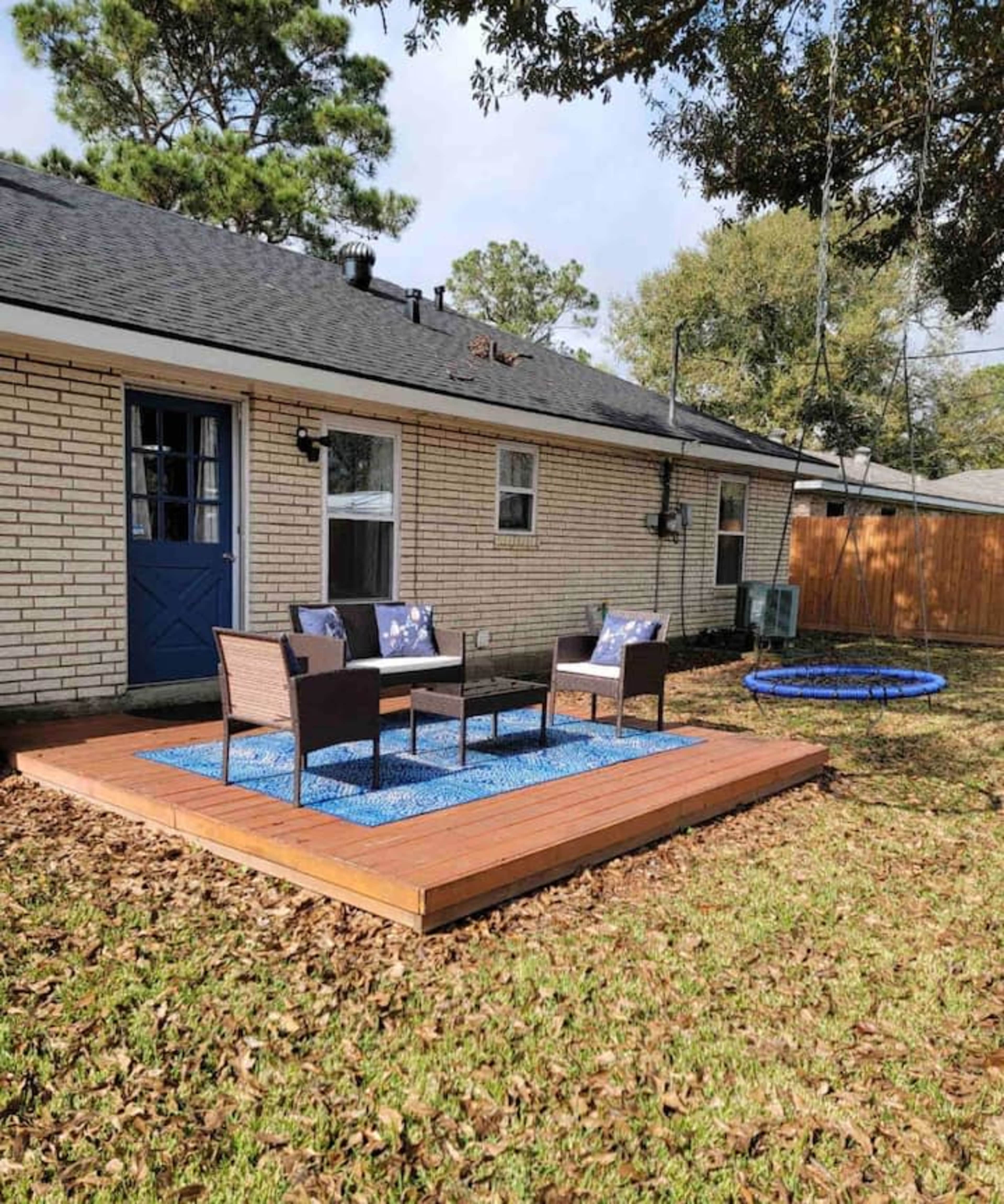 A wooden deck with two chairs and a small table sits in a grassy yard, adjacent to a house with a blue door and a swing hanging from a tree.
