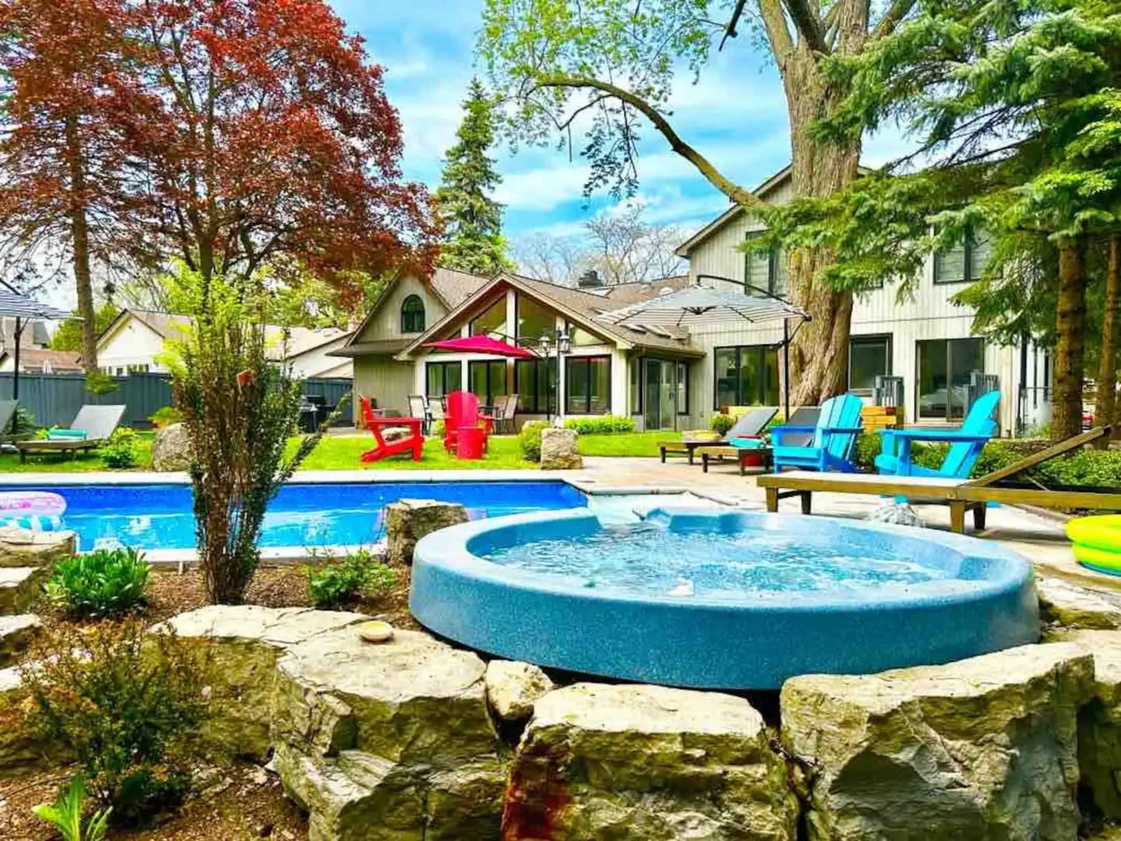 A backyard with a swimming pool, a hot tub made of stone, and colorful lounge chairs surrounded by trees and greenery.