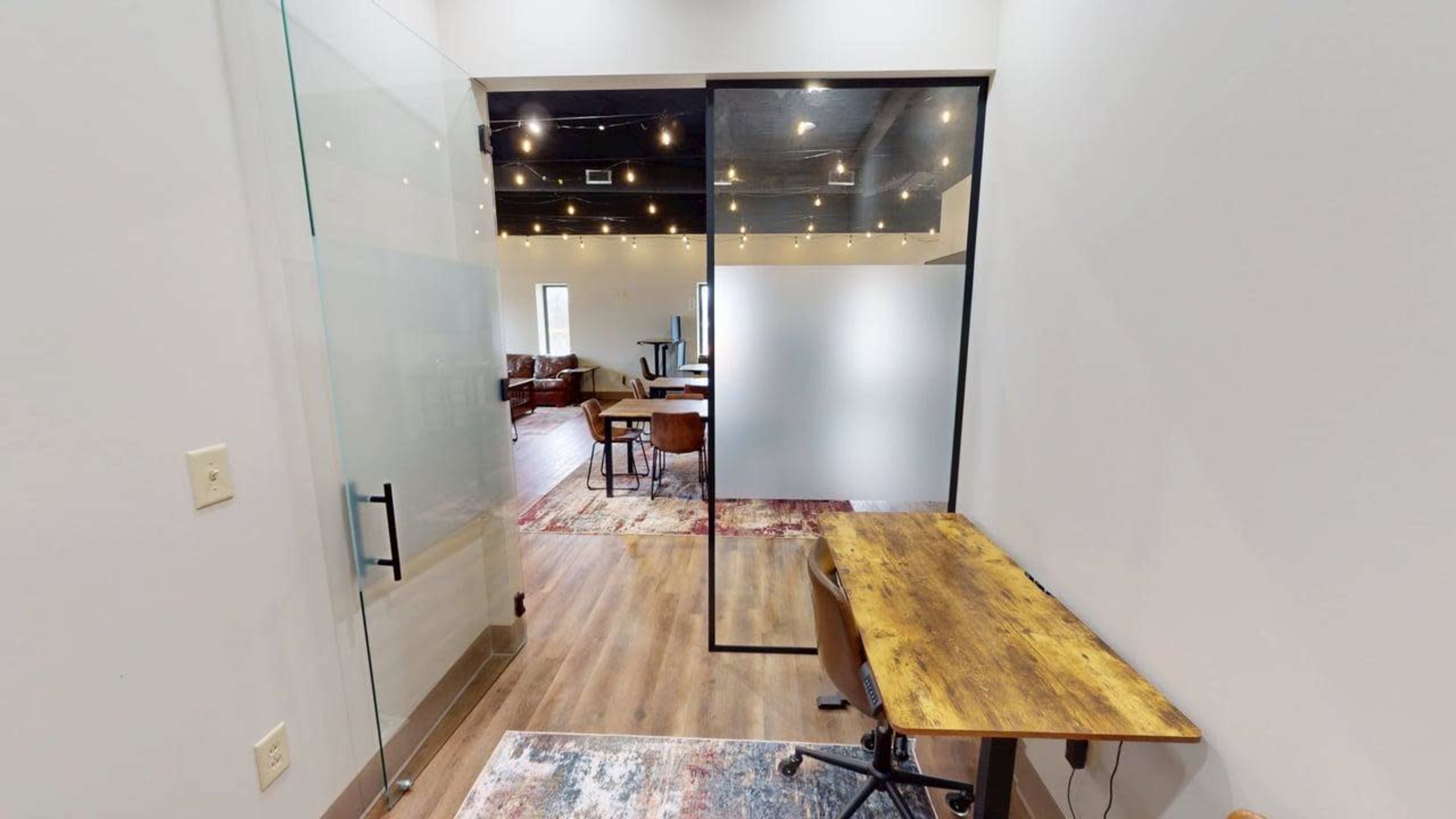The image shows a modern office space featuring a glass-walled meeting room with a wooden desk and a visible lounge area in the background.