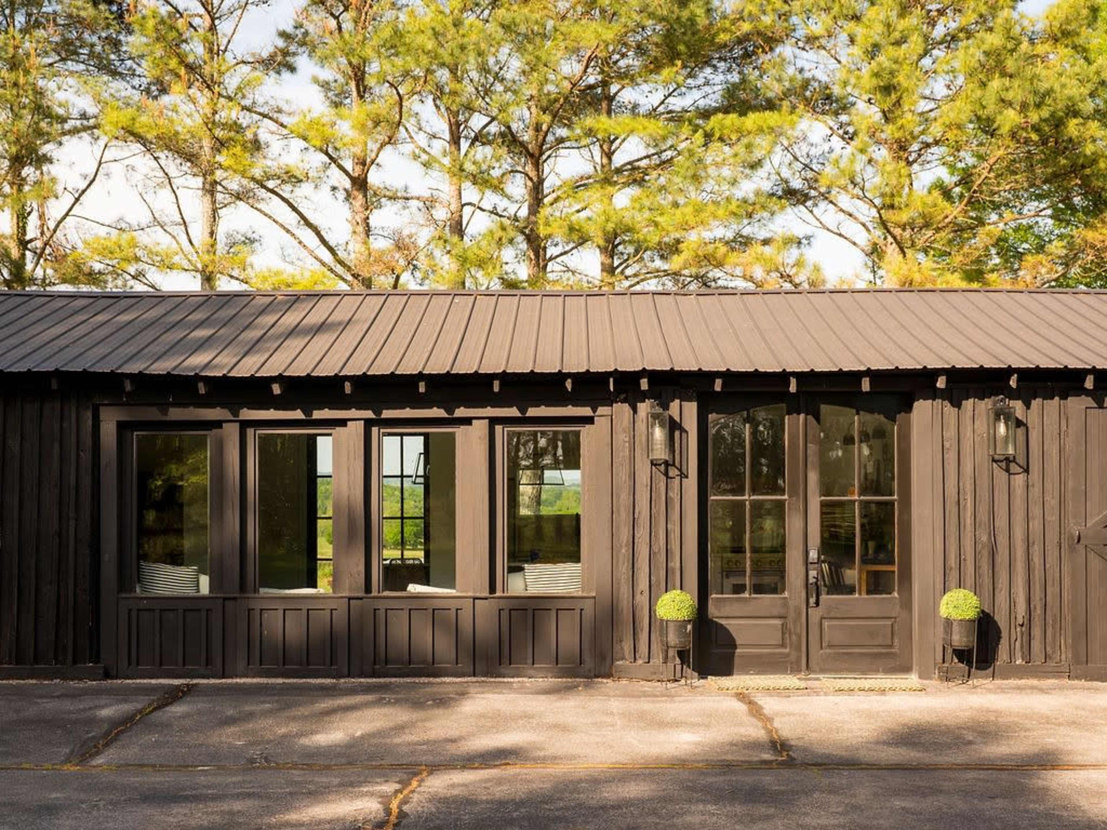 The image shows a black wooden building with large windows and a double door, surrounded by tall trees.
