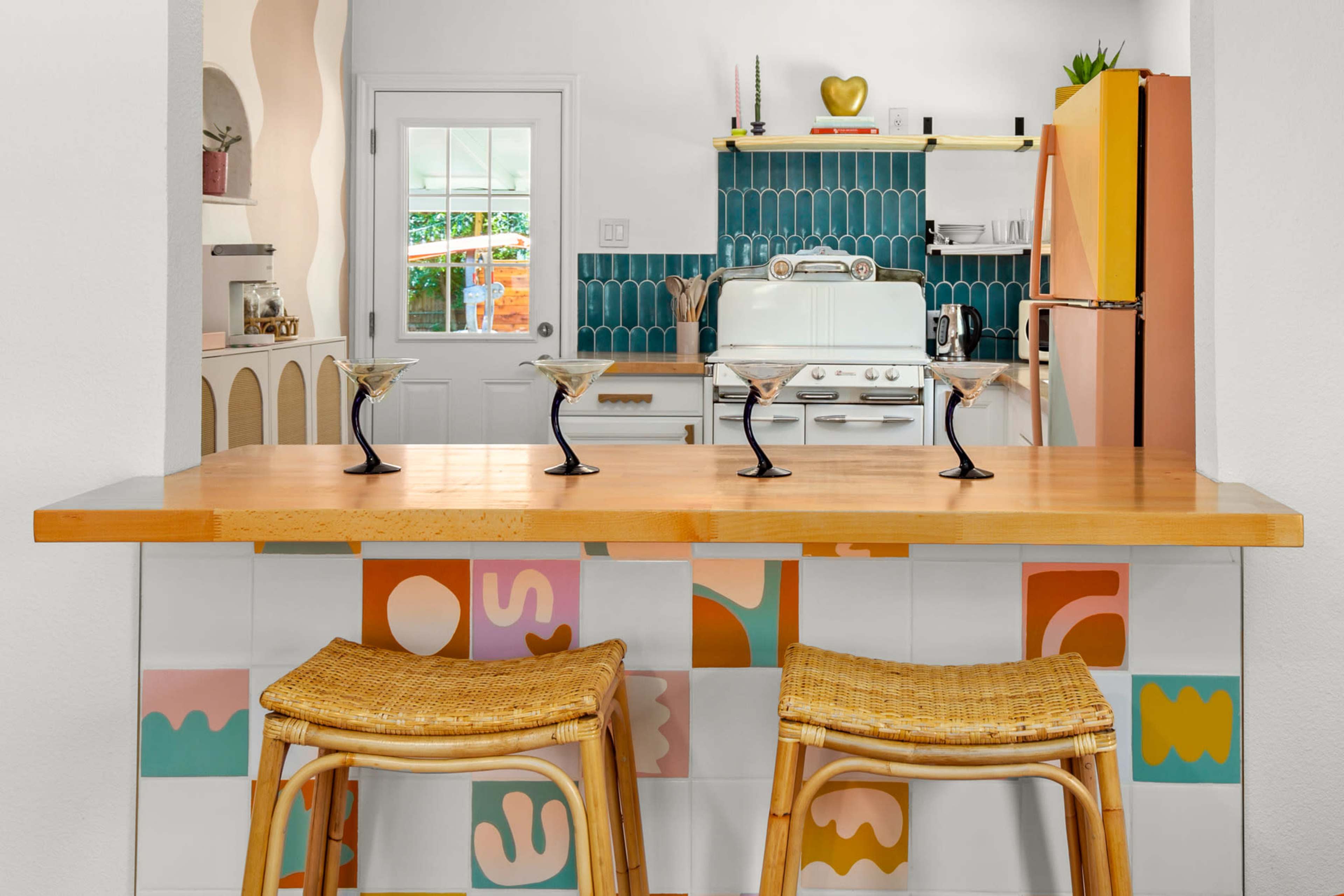 The image features a colorful kitchen with a wooden bar top, two woven stools, and a pastel tile backsplash behind a retro-style stove and refrigerator.