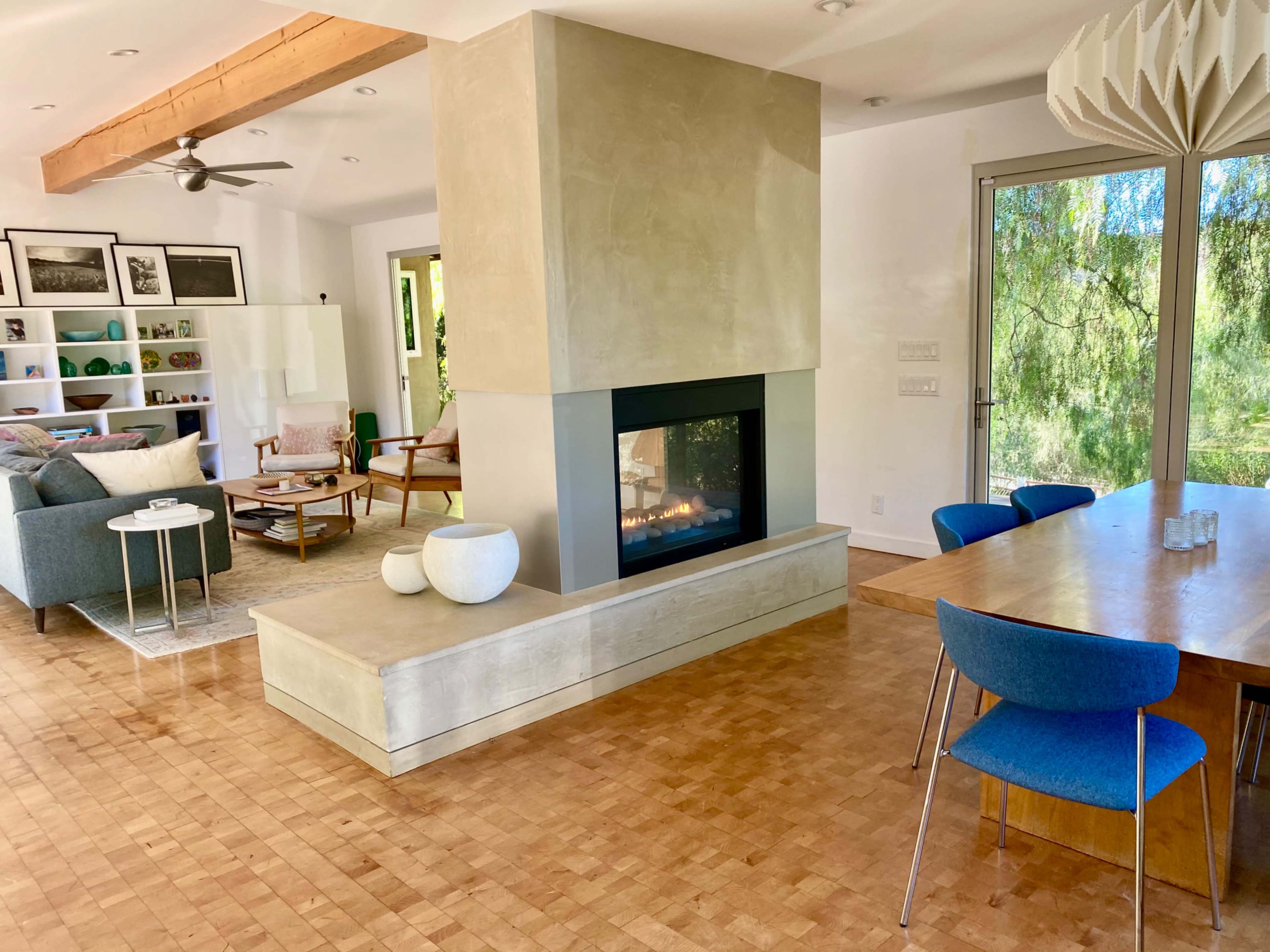 The image shows a modern living space featuring a central fireplace, dining area, and a seating area with large windows overlooking greenery.