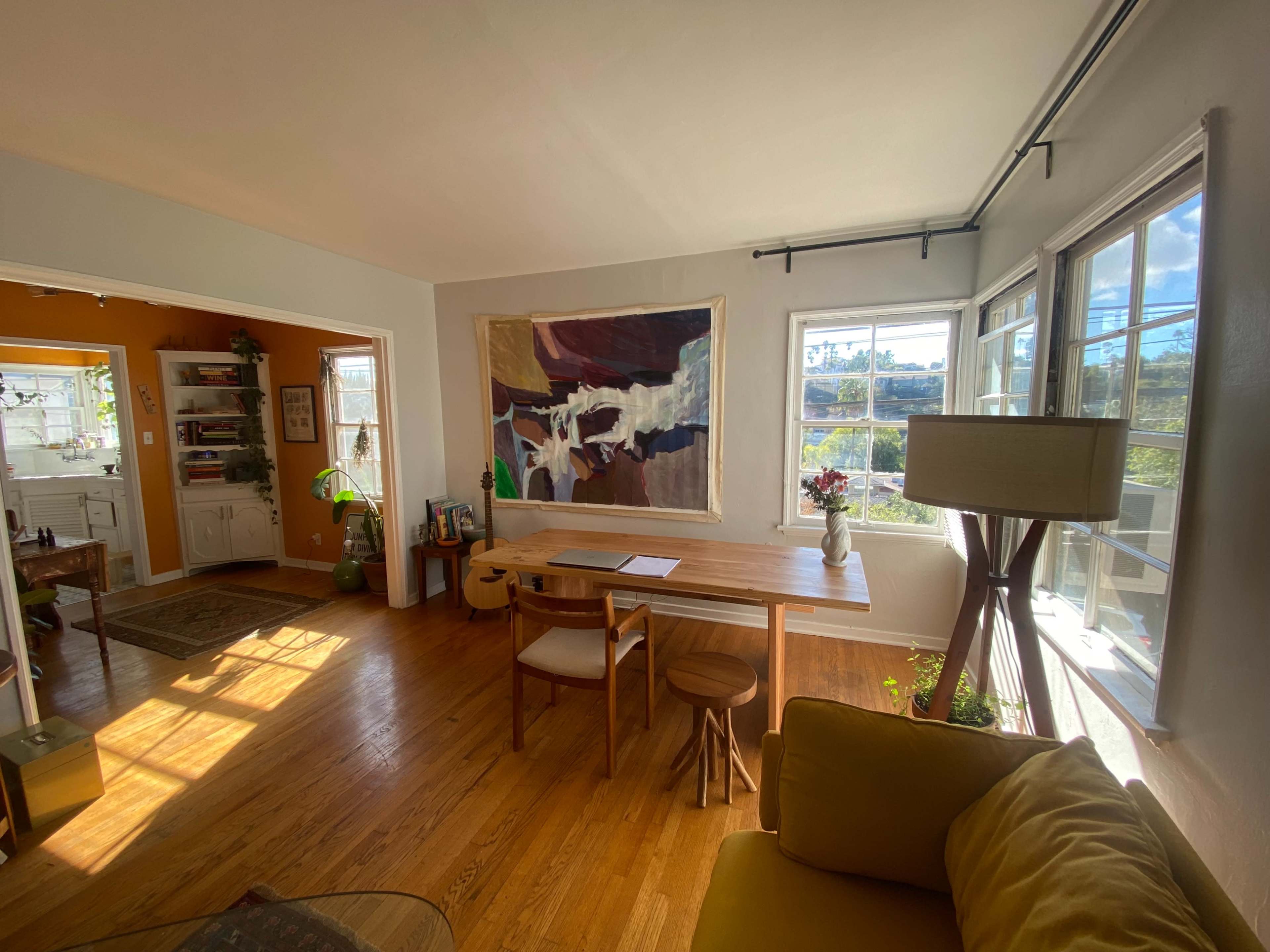 A bright and spacious living area features a wooden dining table with chairs, a large abstract textile art piece on the wall, and large windows allowing natural light to fill the room.