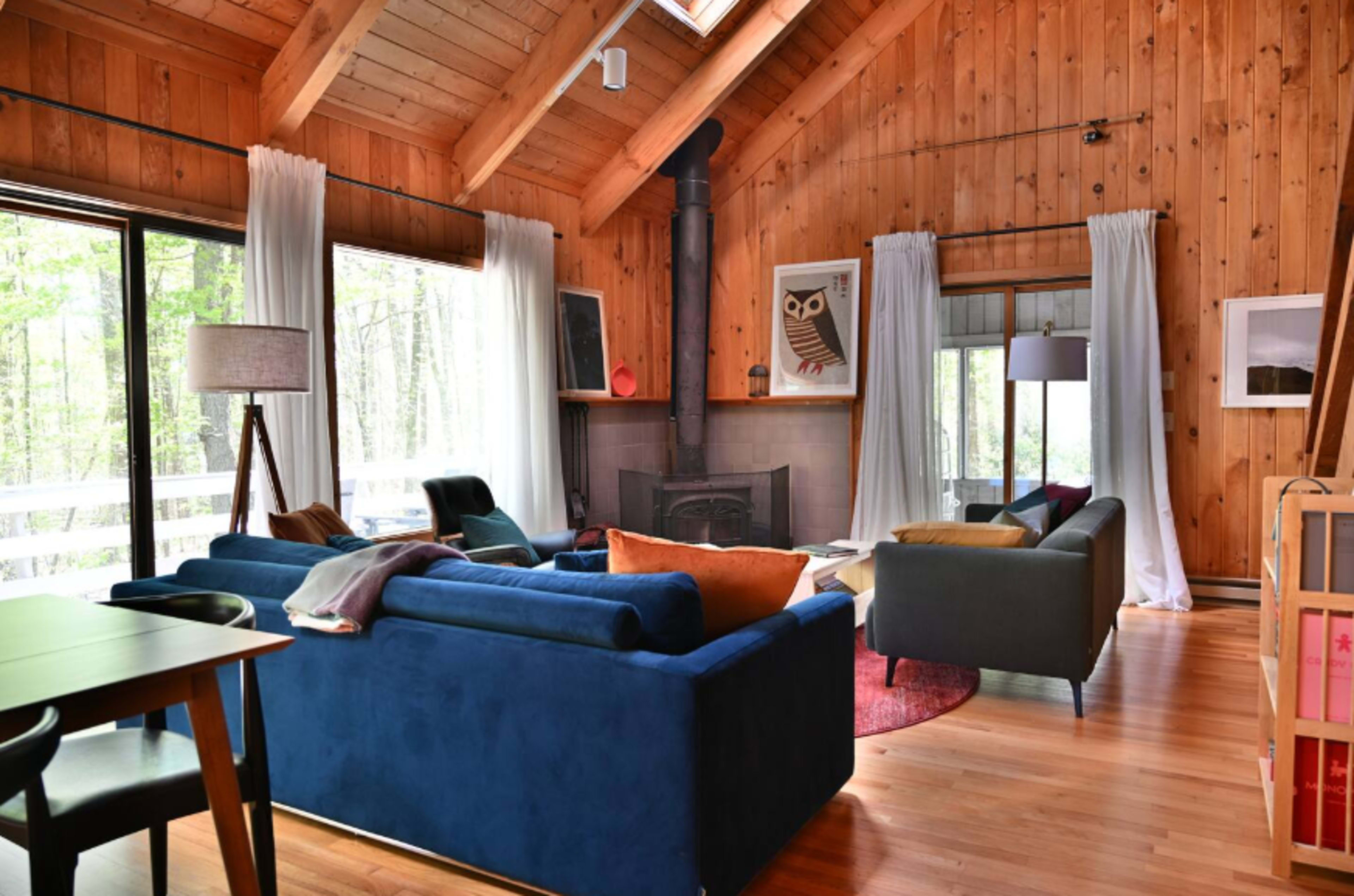 A cozy living room features wooden walls, large windows with natural light, and a mix of blue and green furniture arranged around a central fireplace.