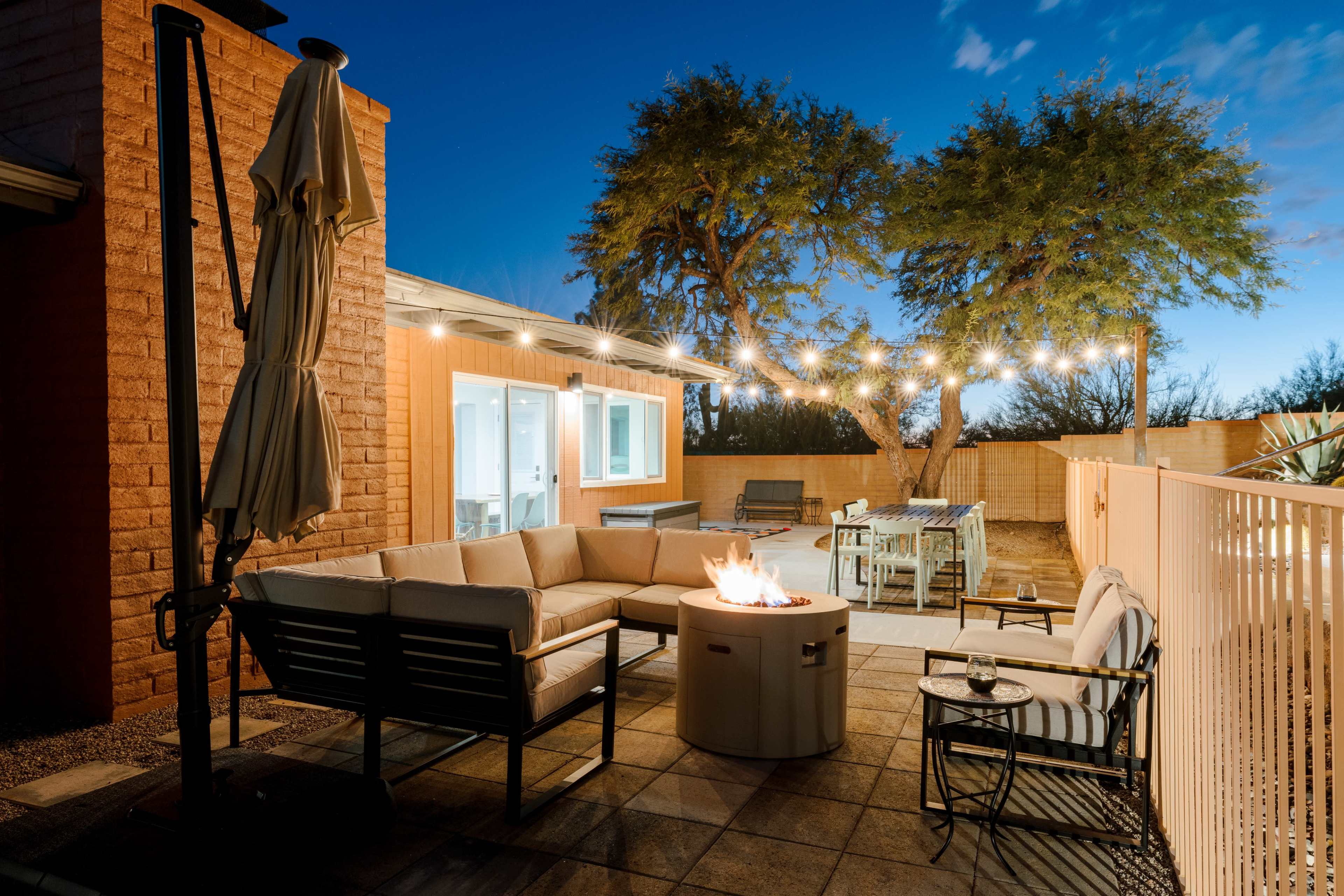 A patio area with a circular fire pit surrounded by cushioned seating, string lights overhead, and a dining table in the background under two trees.