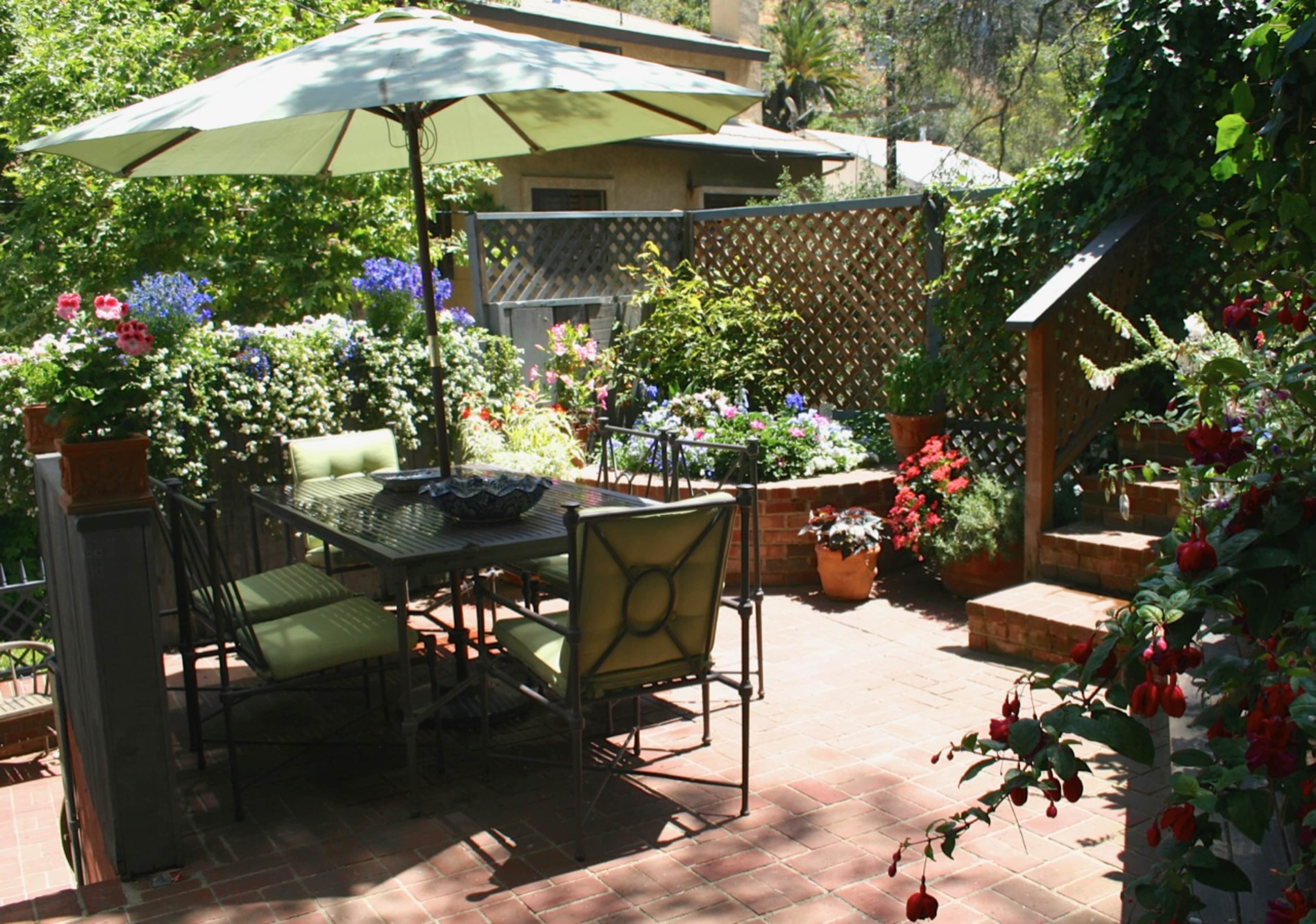 A patio with a dining table and chairs is surrounded by colorful flower pots and plants, with an umbrella providing shade.