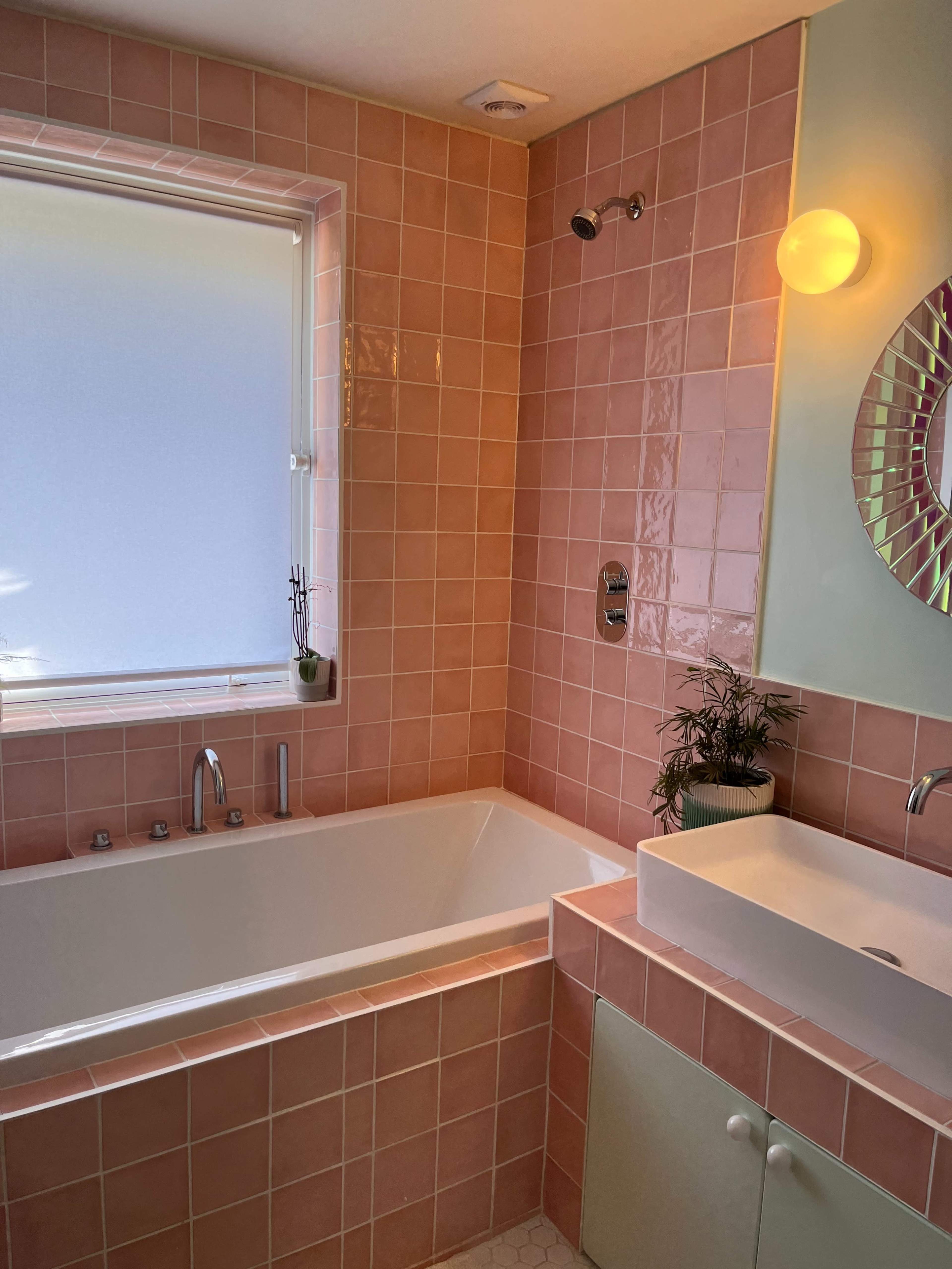 A bathroom with pink tiled walls, a white bathtub, a modern sink, and a window letting in natural light.