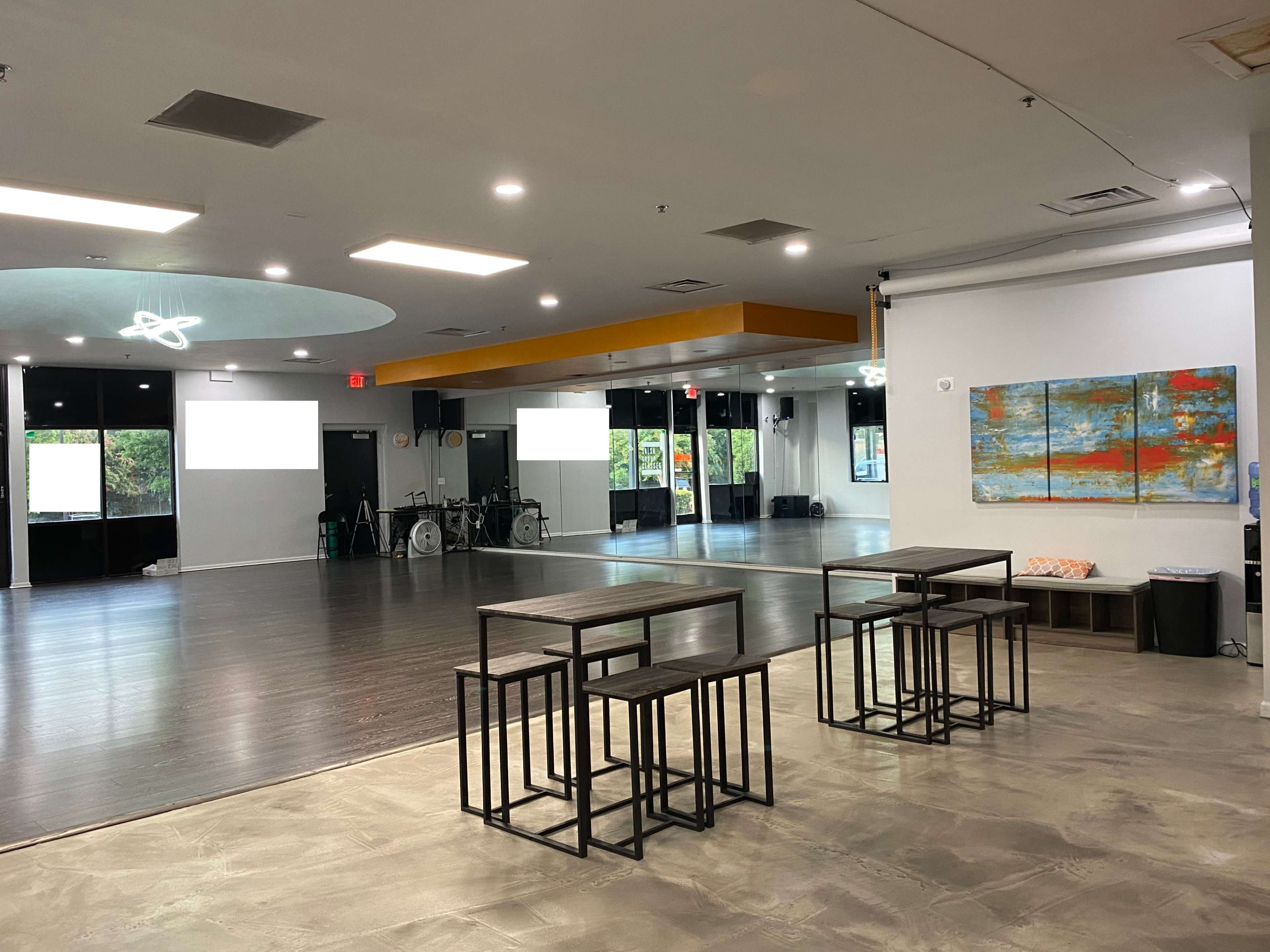 A spacious studio with a polished wooden floor, two sets of square tables in the foreground, and large windows letting in natural light.