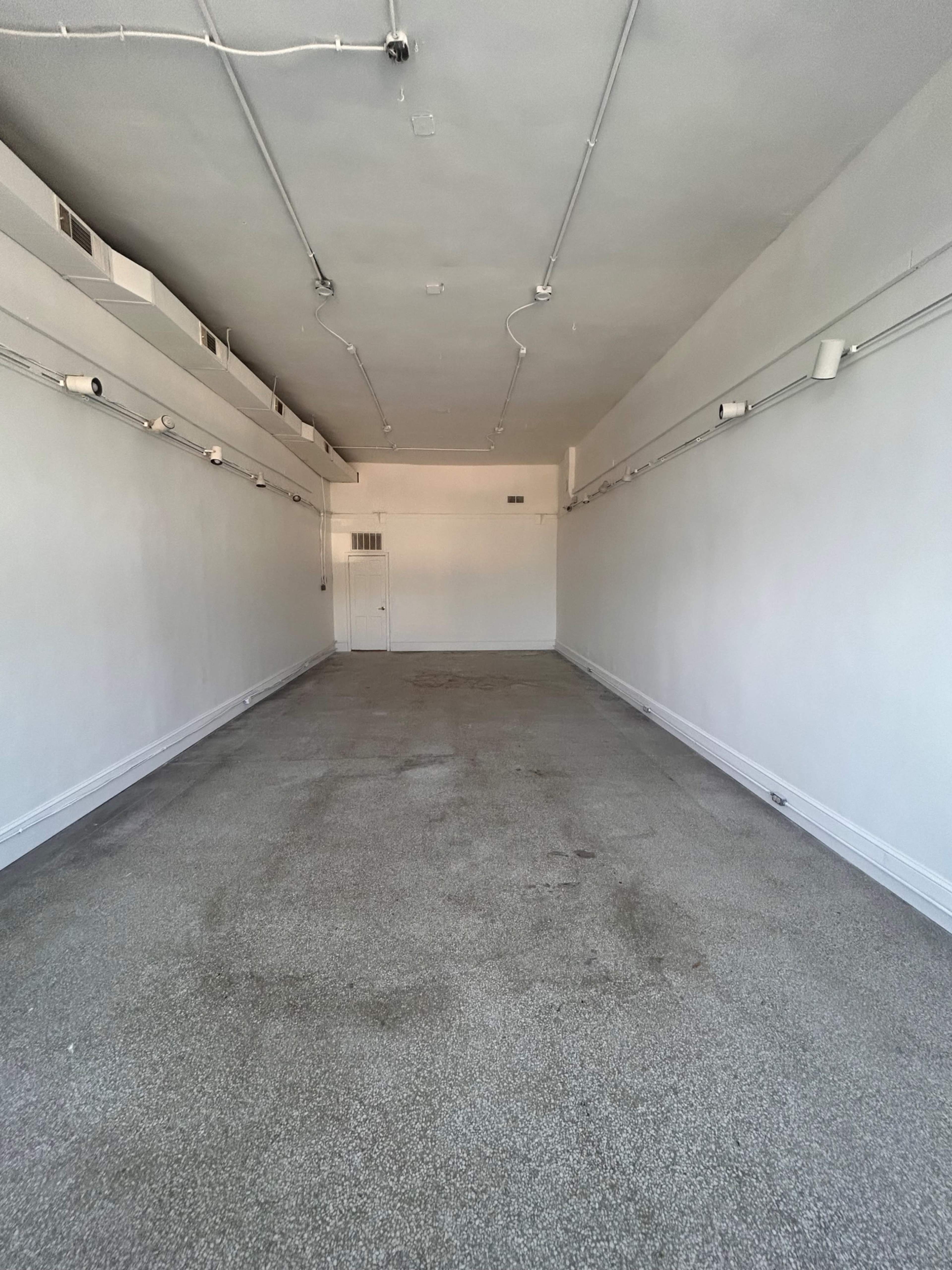 The image shows a spacious, empty room with white walls and a carpeted floor, illuminated by overhead lights.