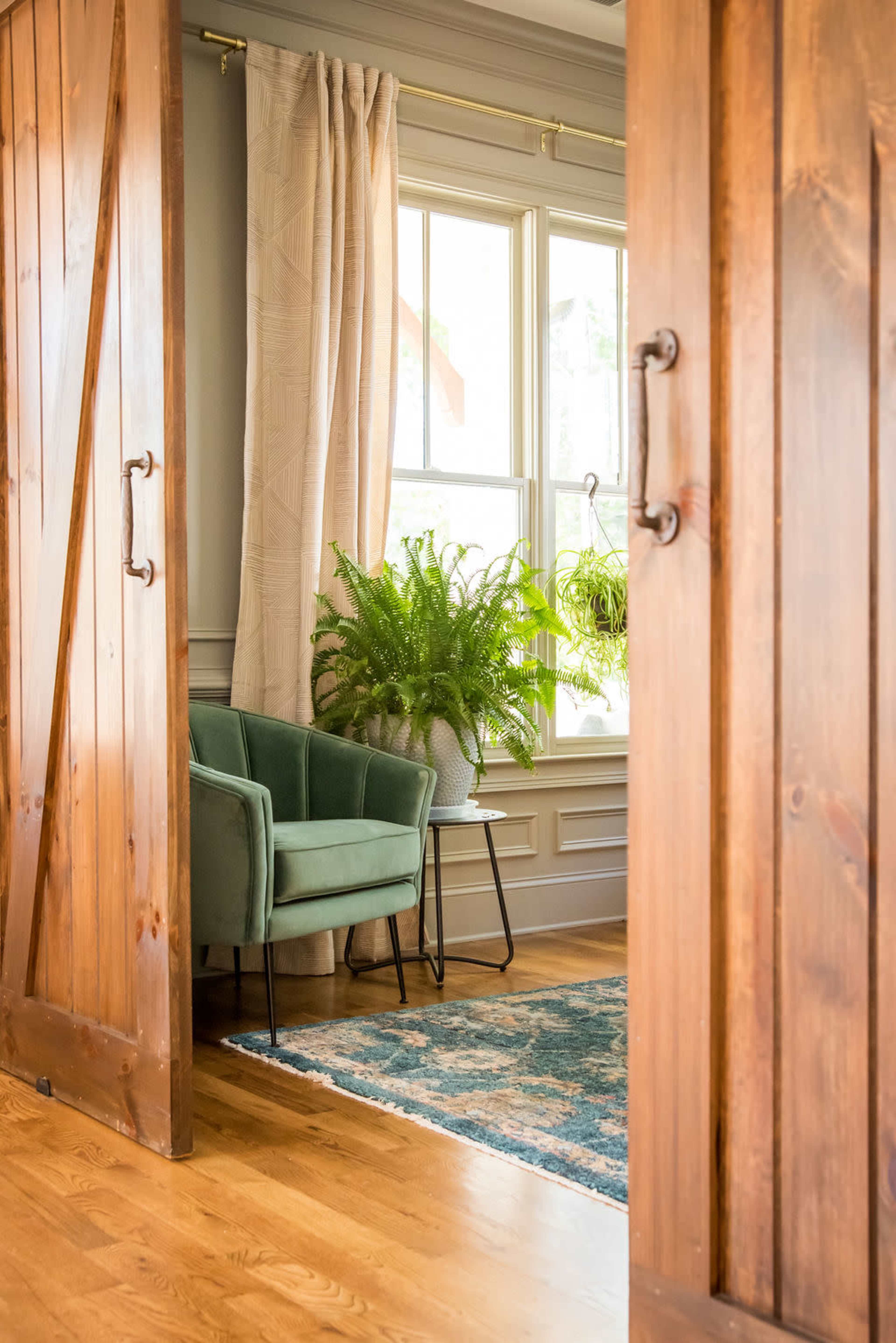 A green chair is positioned next to a small table and a potted fern in a well-lit room visible through open wooden doors.