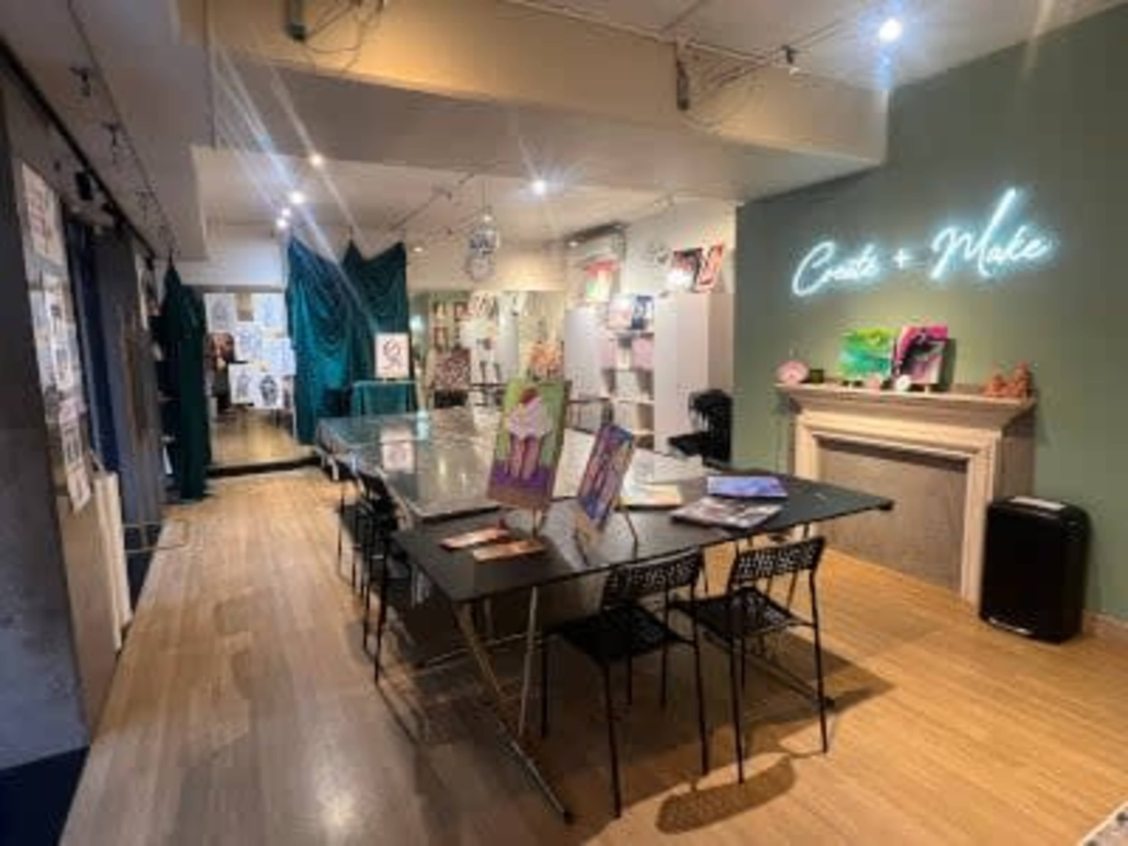 The image shows a spacious creative studio with a large table, black chairs, art supplies, and decorative artwork displayed on the walls.