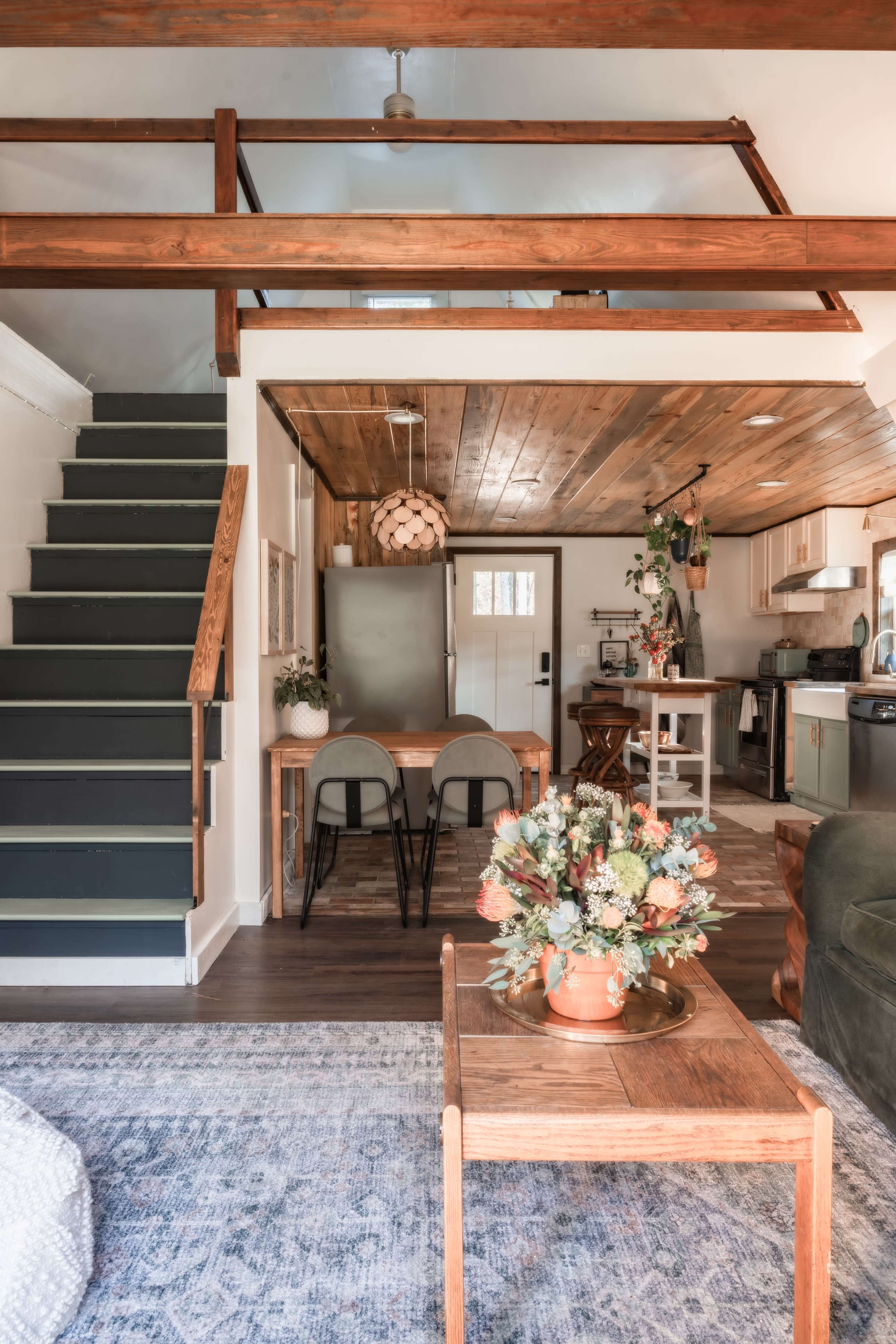 A modern, open-concept living space featuring a dining area, kitchen, and a staircase leading to a lofted area, with a wooden ceiling and floral decor on a coffee table.