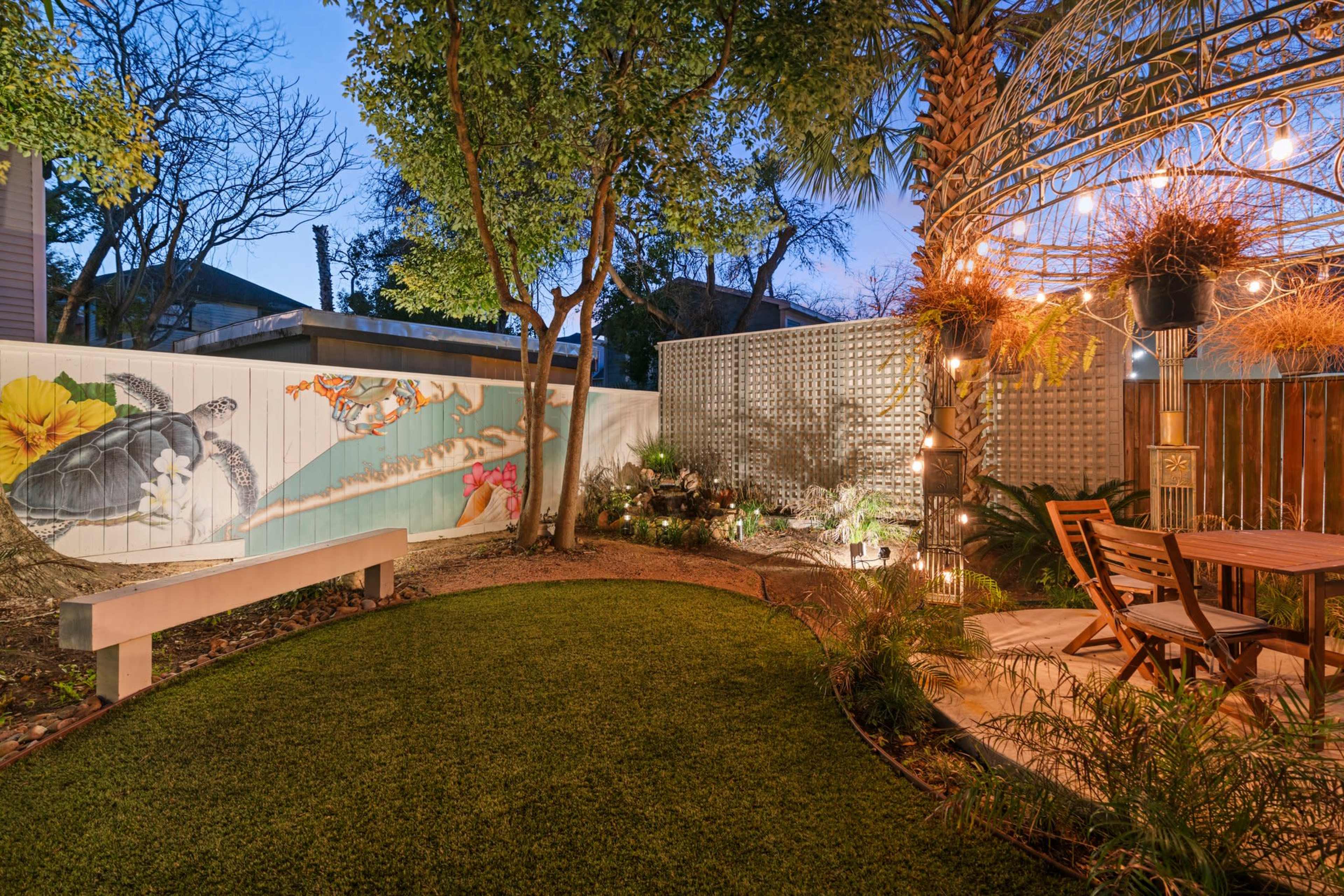 A landscaped garden features a mural of a turtle on a fence, a seating area with a wooden table and chairs, and decorative lighting around a circular structure.