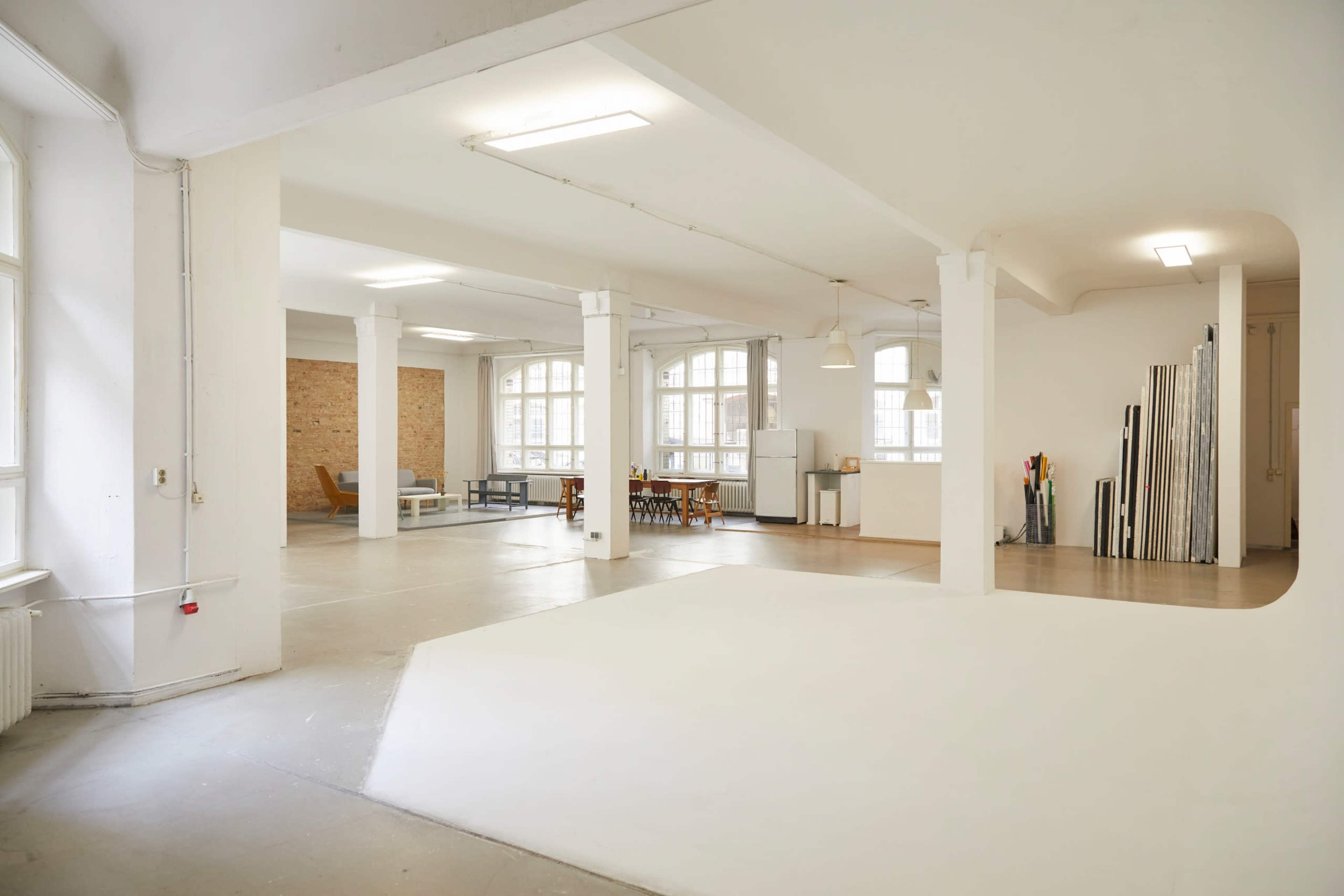 A spacious, well-lit interior of a minimalist studio with white walls, tall windows, and a mixture of furniture and decor items.
