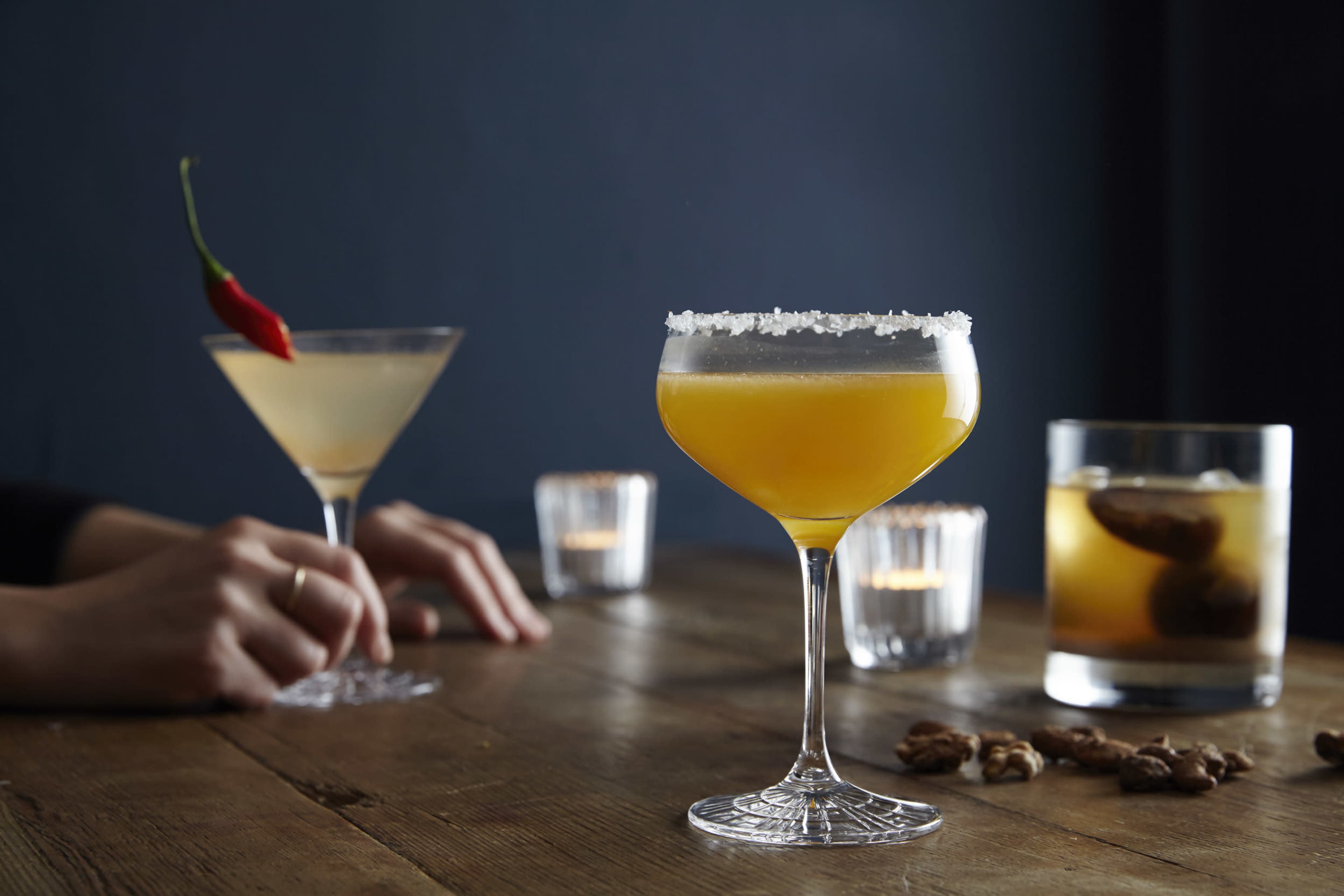 Two cocktails are presented on a wooden table, with a yellow drink garnished with salt and accompanied by another cocktail, while candles illuminate the scene.
