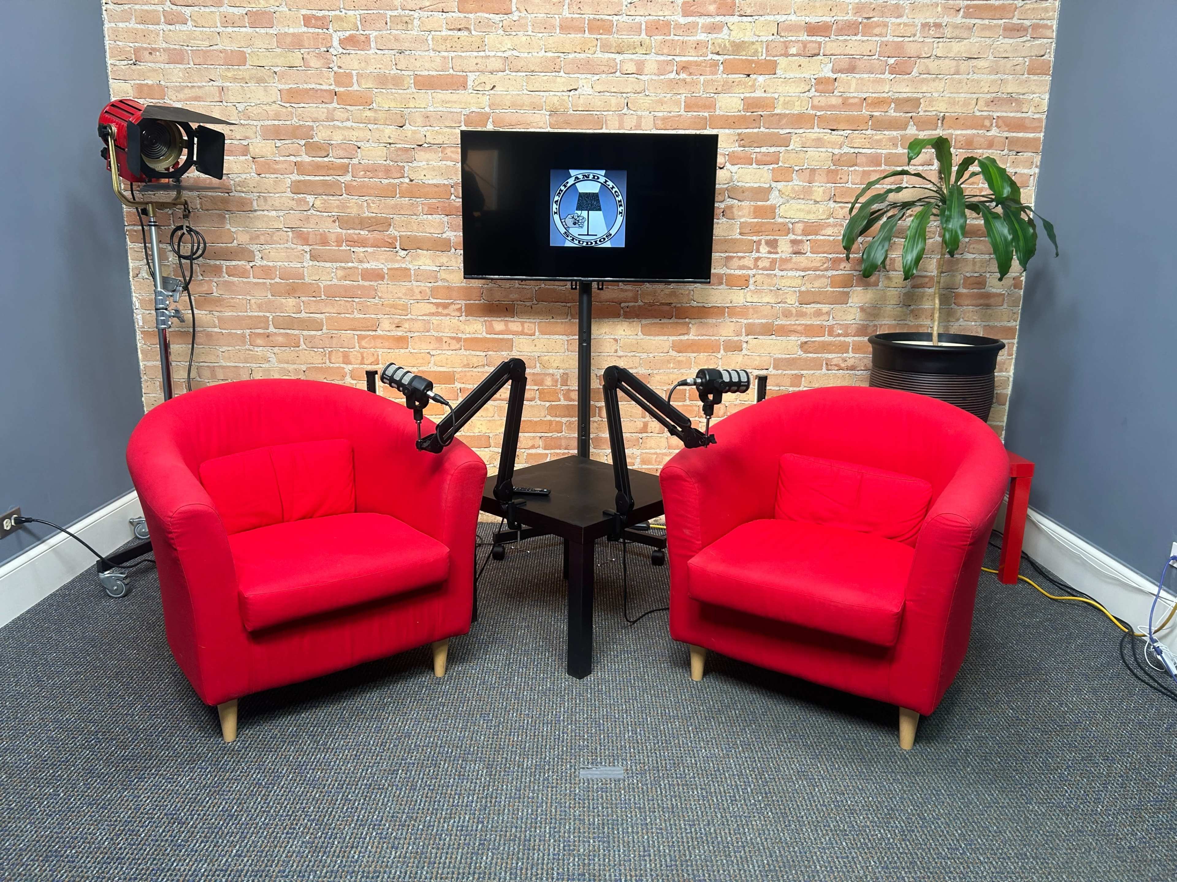 The image shows a podcast studio setup featuring two red chairs, a table in between, a television screen on a stand, and a potted plant against a brick wall.