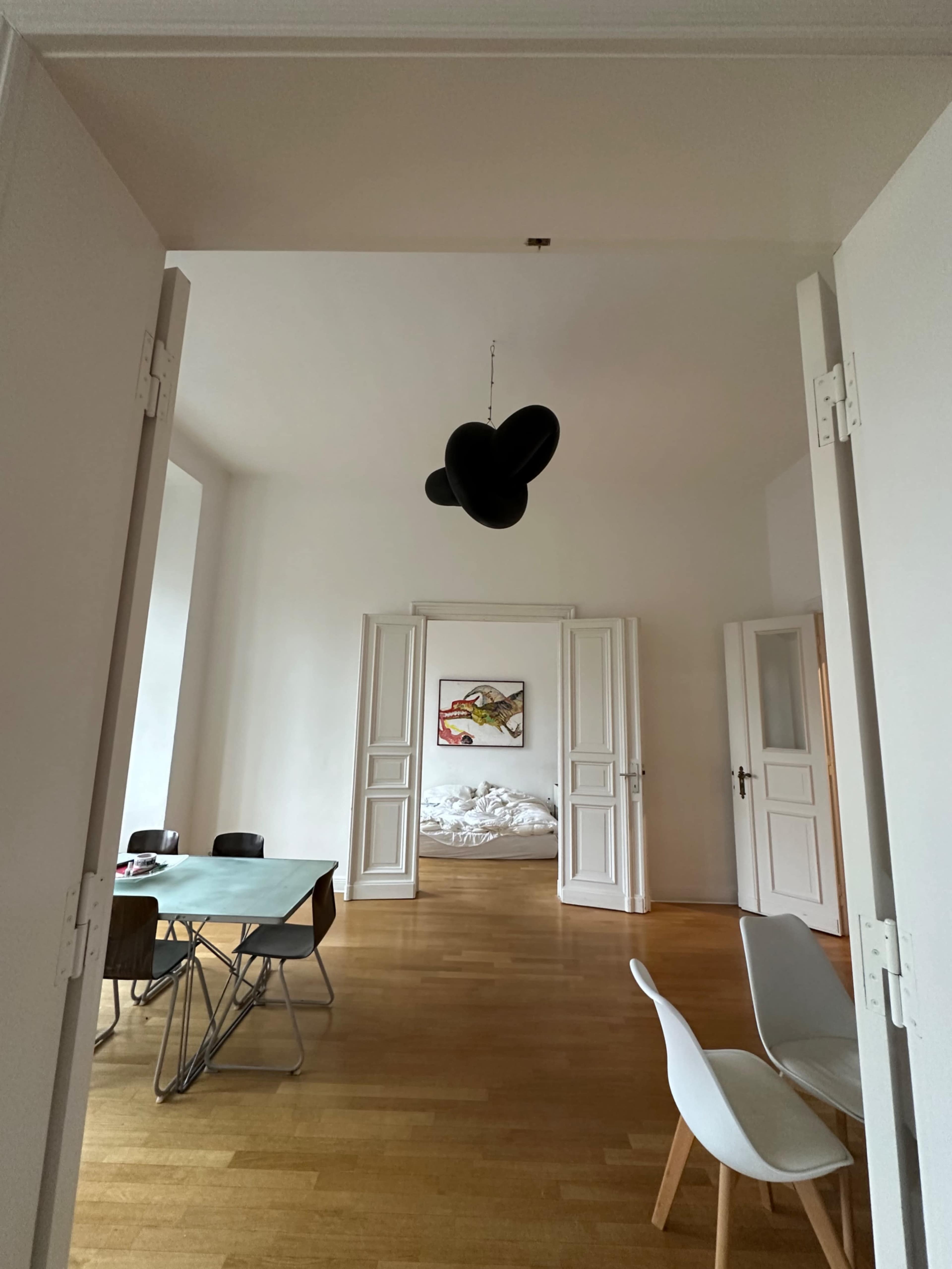 A black pendant light hangs in an open room leading to a bedroom with a disheveled bed, while a dining table is set with chairs on the left.