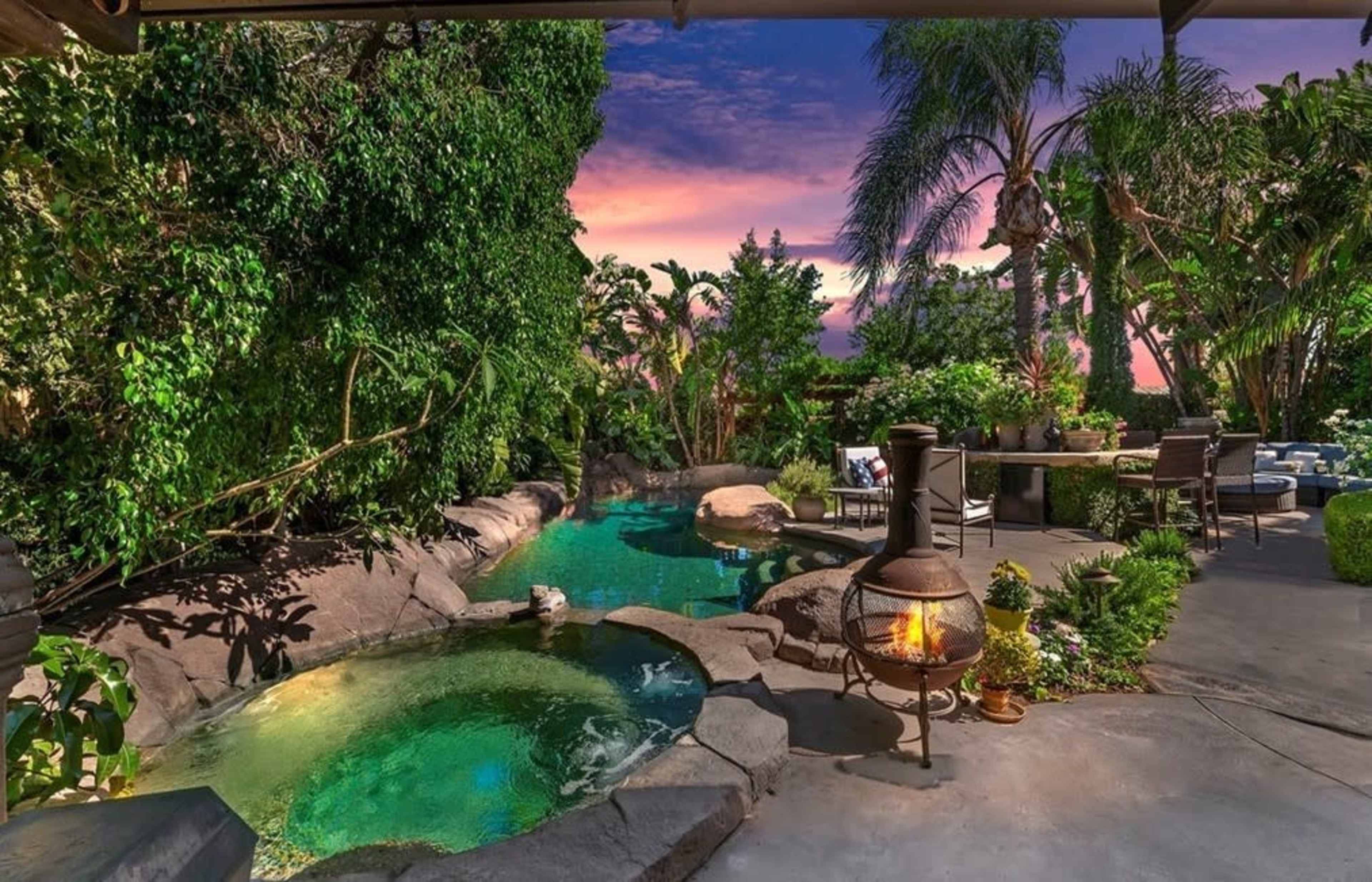 A landscaped backyard featuring a stone pool, a hot tub, and a fire pit surrounded by lush greenery at sunset.