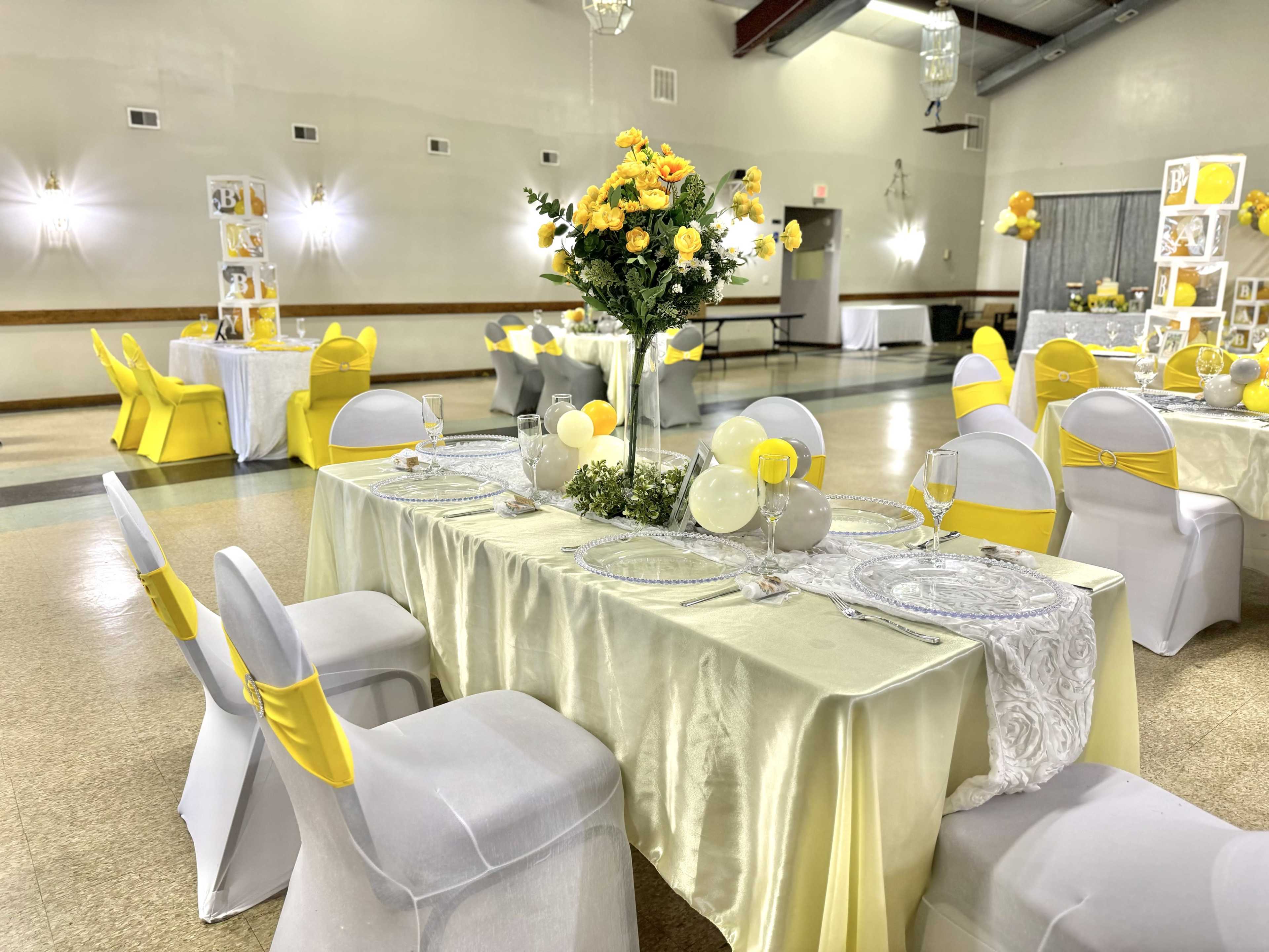 A banquet hall is set up with tables dressed in light yellow tablecloths, adorned with floral centerpieces and decorated with yellow accents, while chairs are covered in white with yellow sashes.