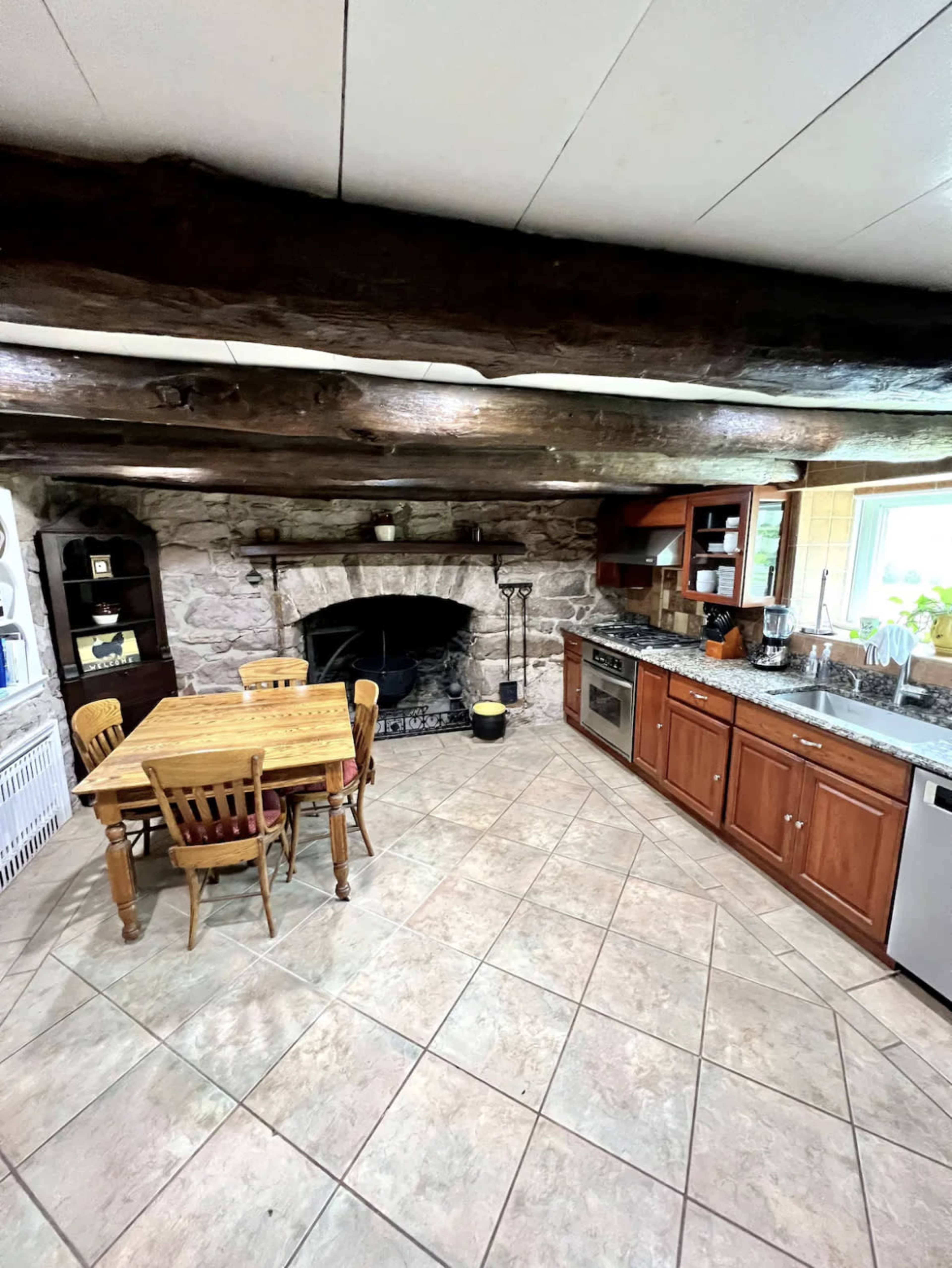 The kitchen features a stone fireplace, wooden beams, a tiled floor, and a dining table surrounded by chairs.
