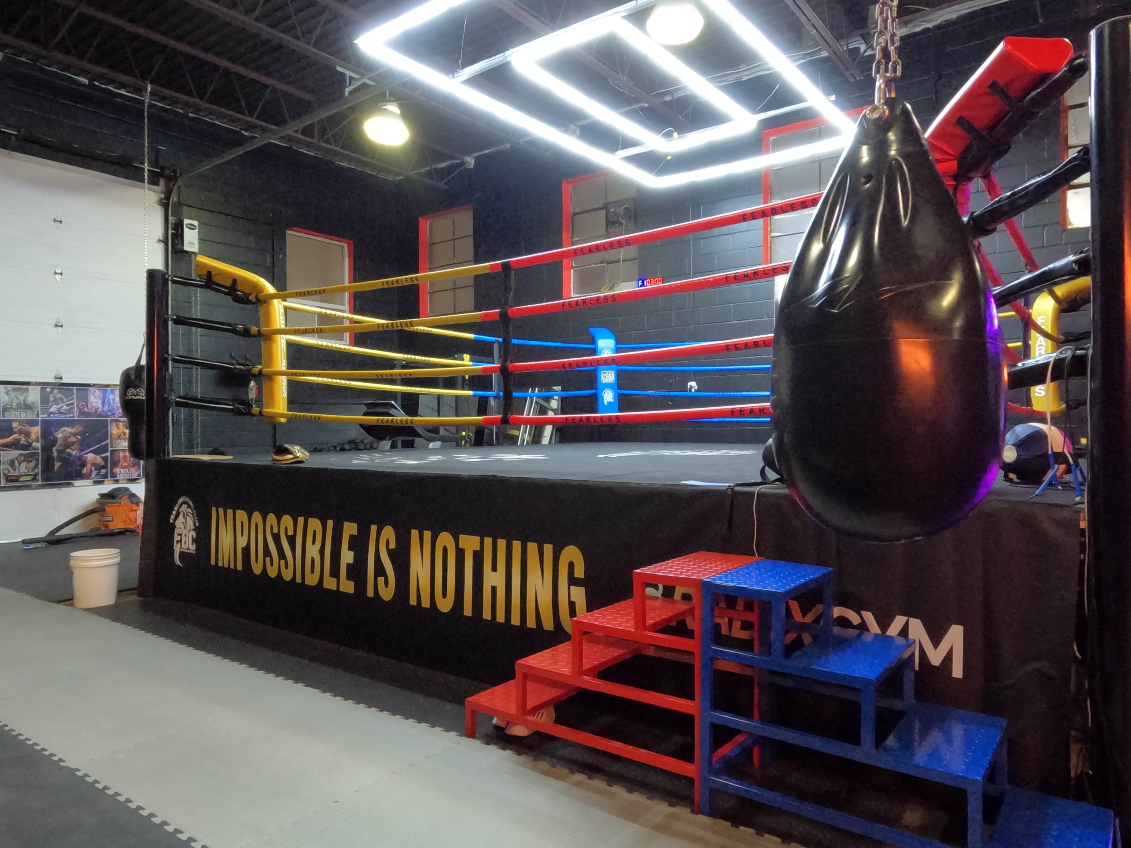 The image shows a boxing gym with a red and yellow ring, a punching bag hanging nearby, and a staircase leading up to the ring.