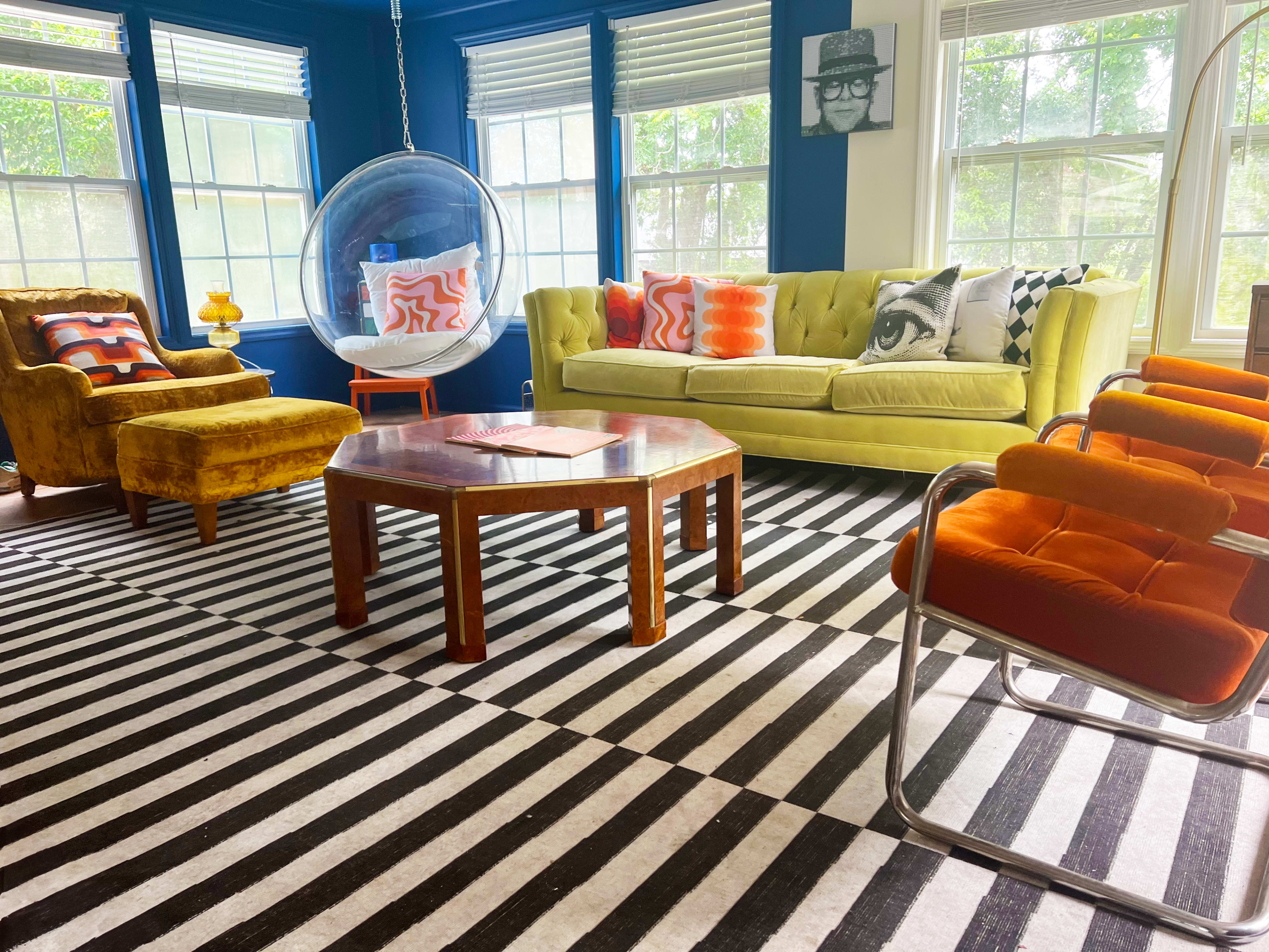 The room features a bright green sofa, an orange accent chair, a black and white striped rug, and a round glass bubble chair, creating a vibrant living space.