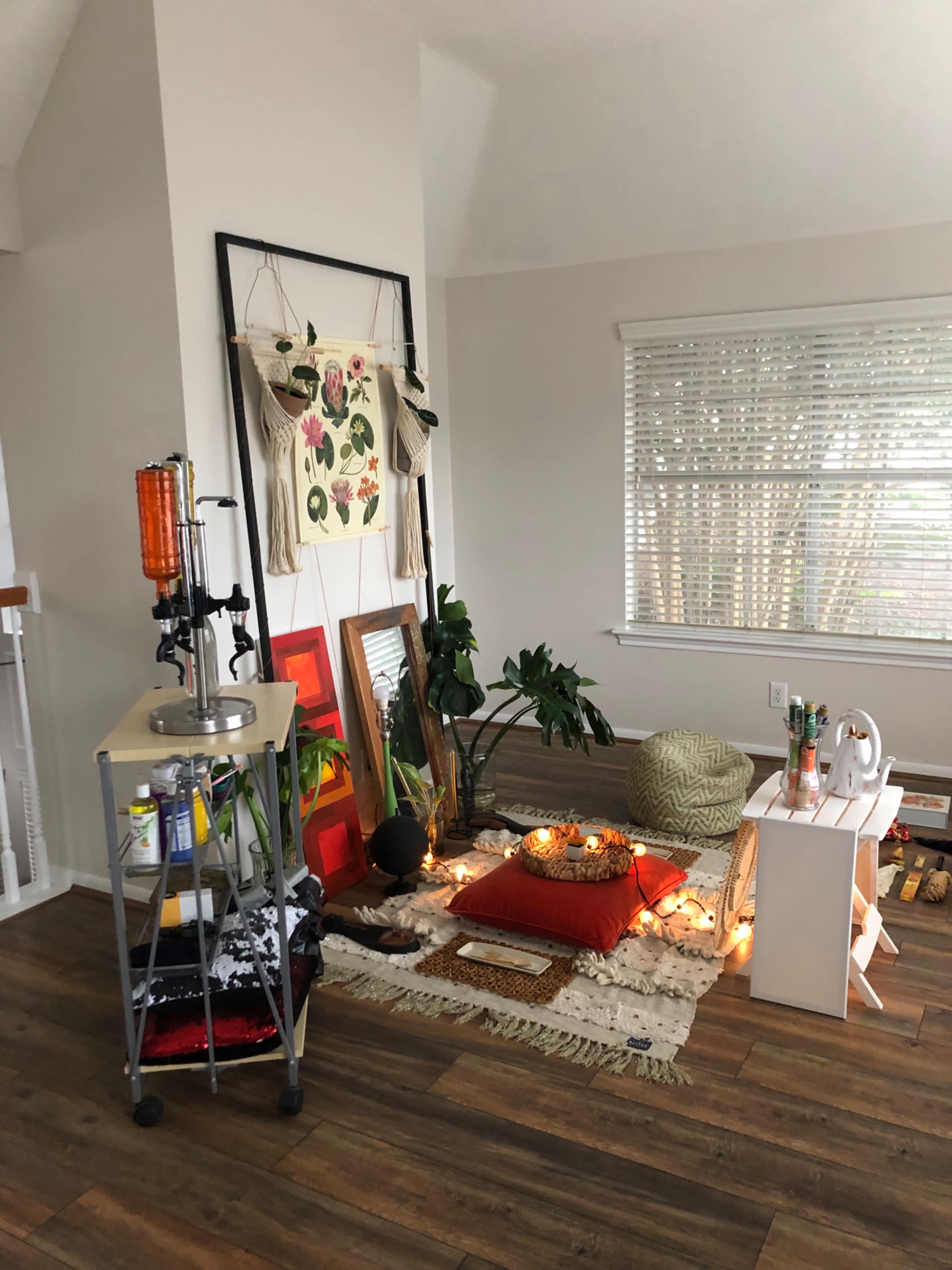 A cozy corner of a room features a relaxed seating area with cushions, plants, and decorative items, illuminated by string lights.