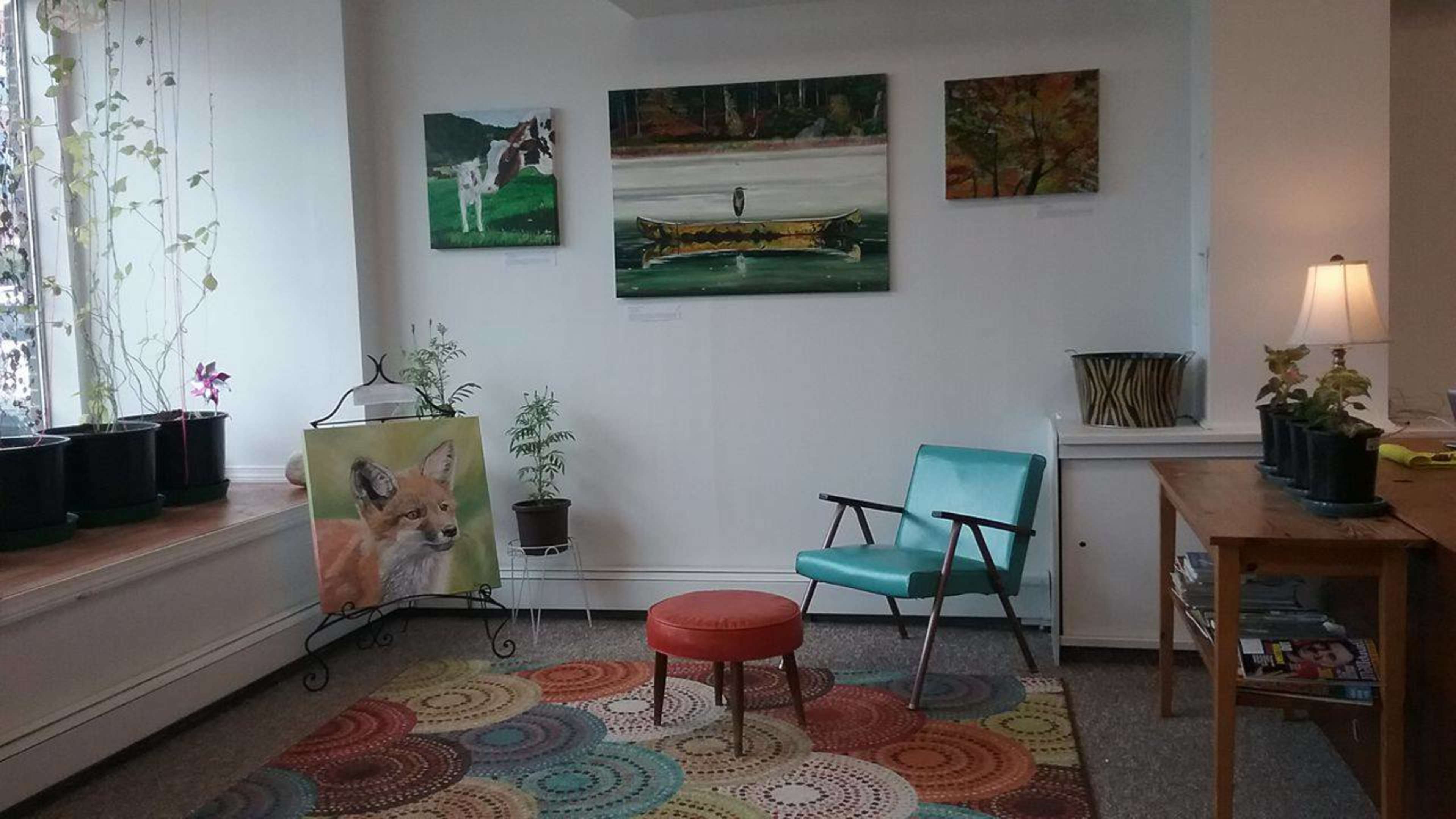 The room features a brightly colored rug, an aqua chair, a small red stool, potted plants, and several paintings on the walls.