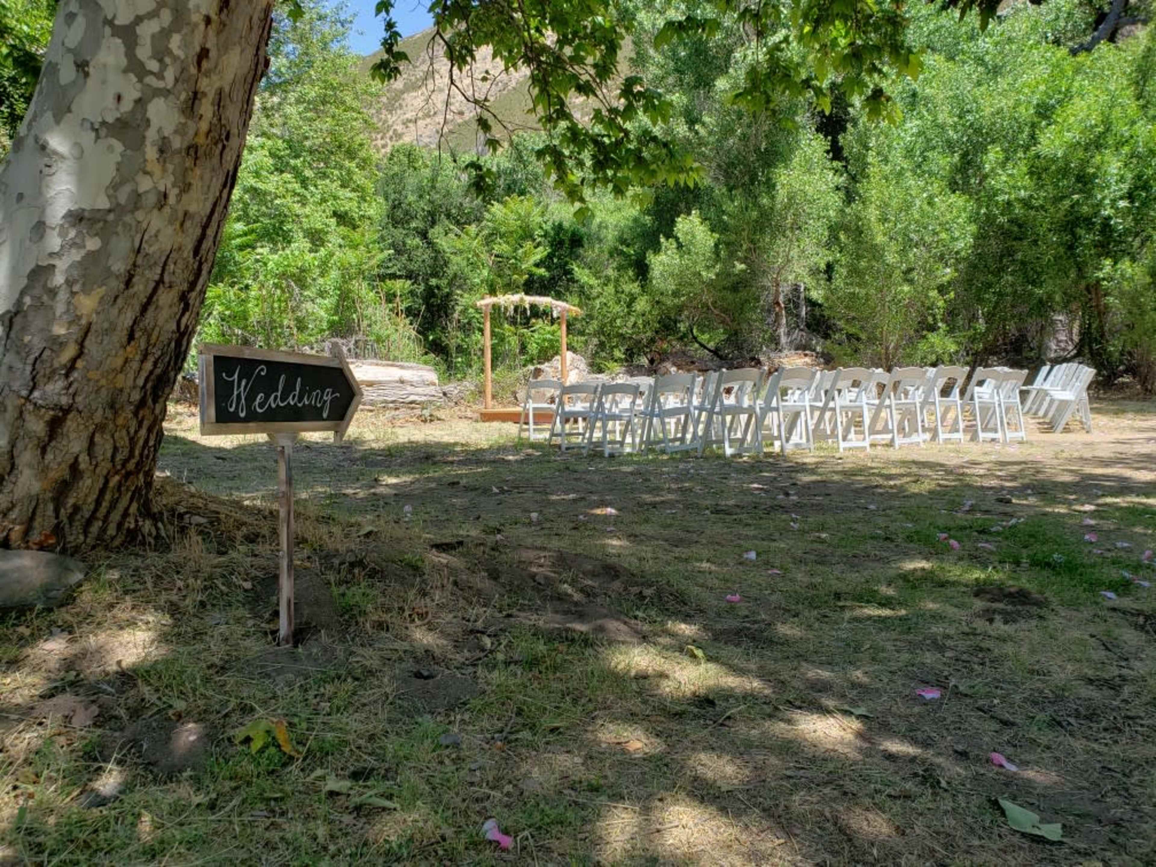 A rustic wedding setup features rows of white chairs arranged near a wooden arch under a tree.