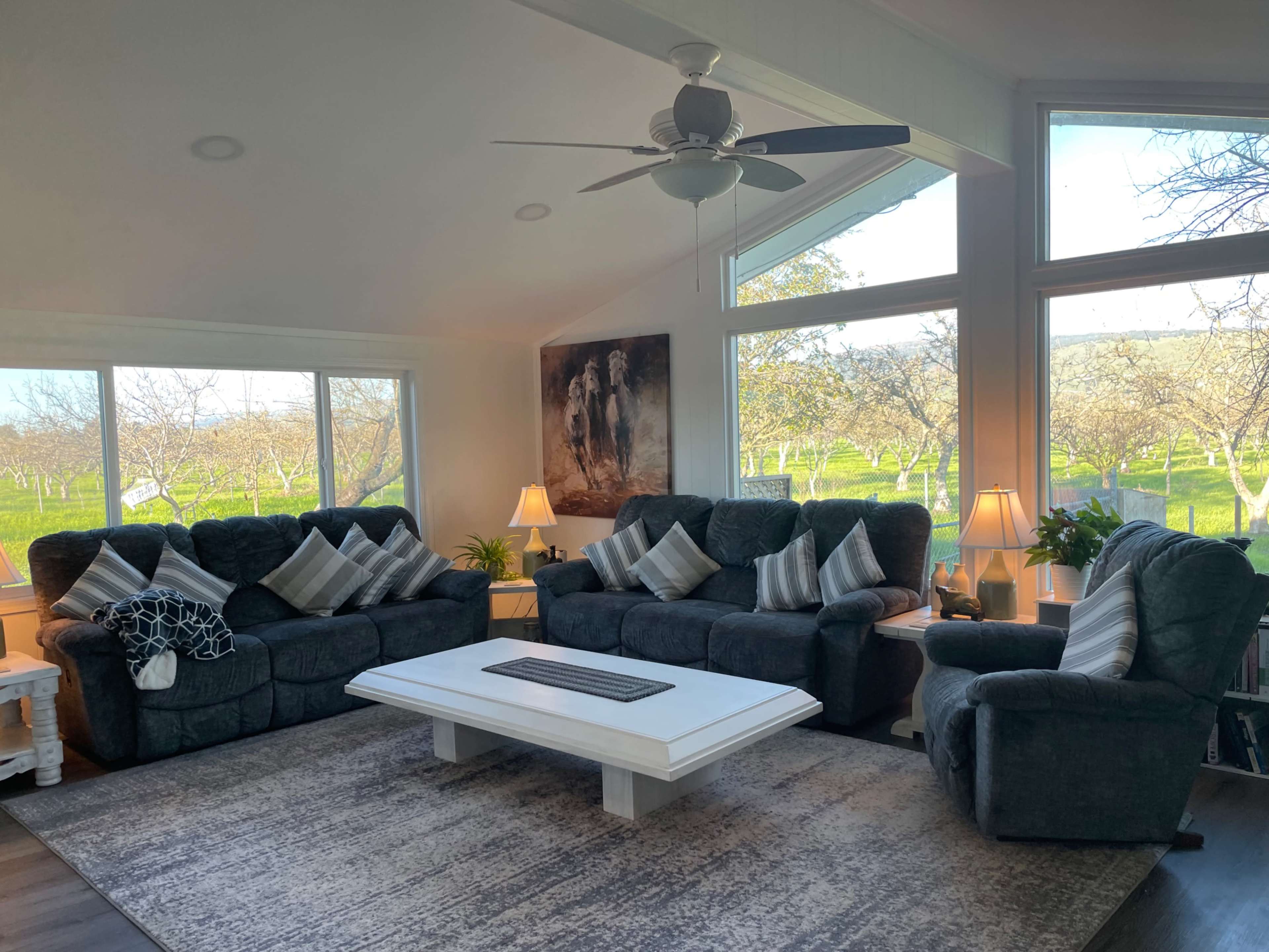 A modern living room features two dark sofas and a coffee table, with large windows overlooking an orchard.
