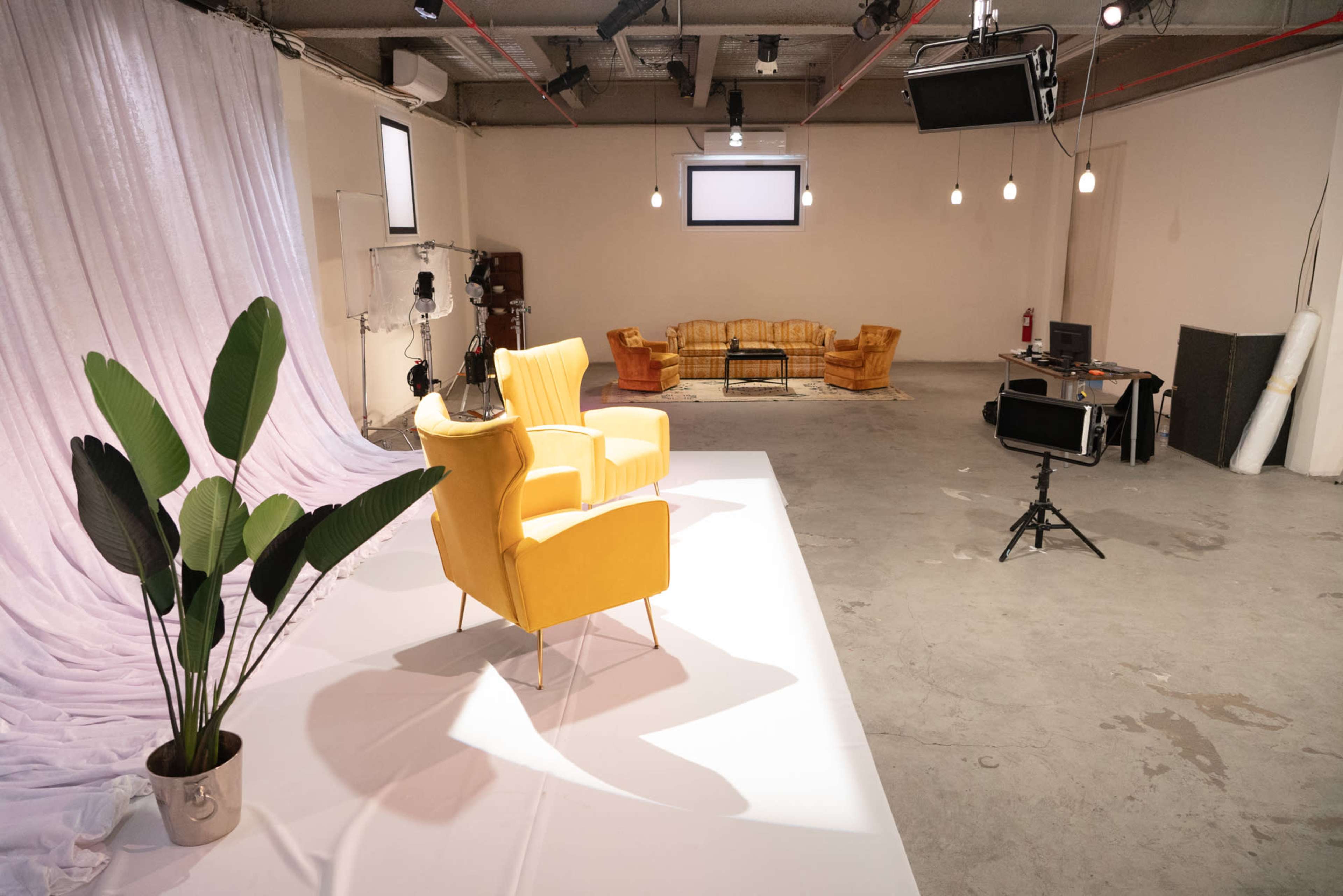 A brightly lit studio features a yellow armchair on a white stage, accompanied by a potted plant and two yellow couches in the background.