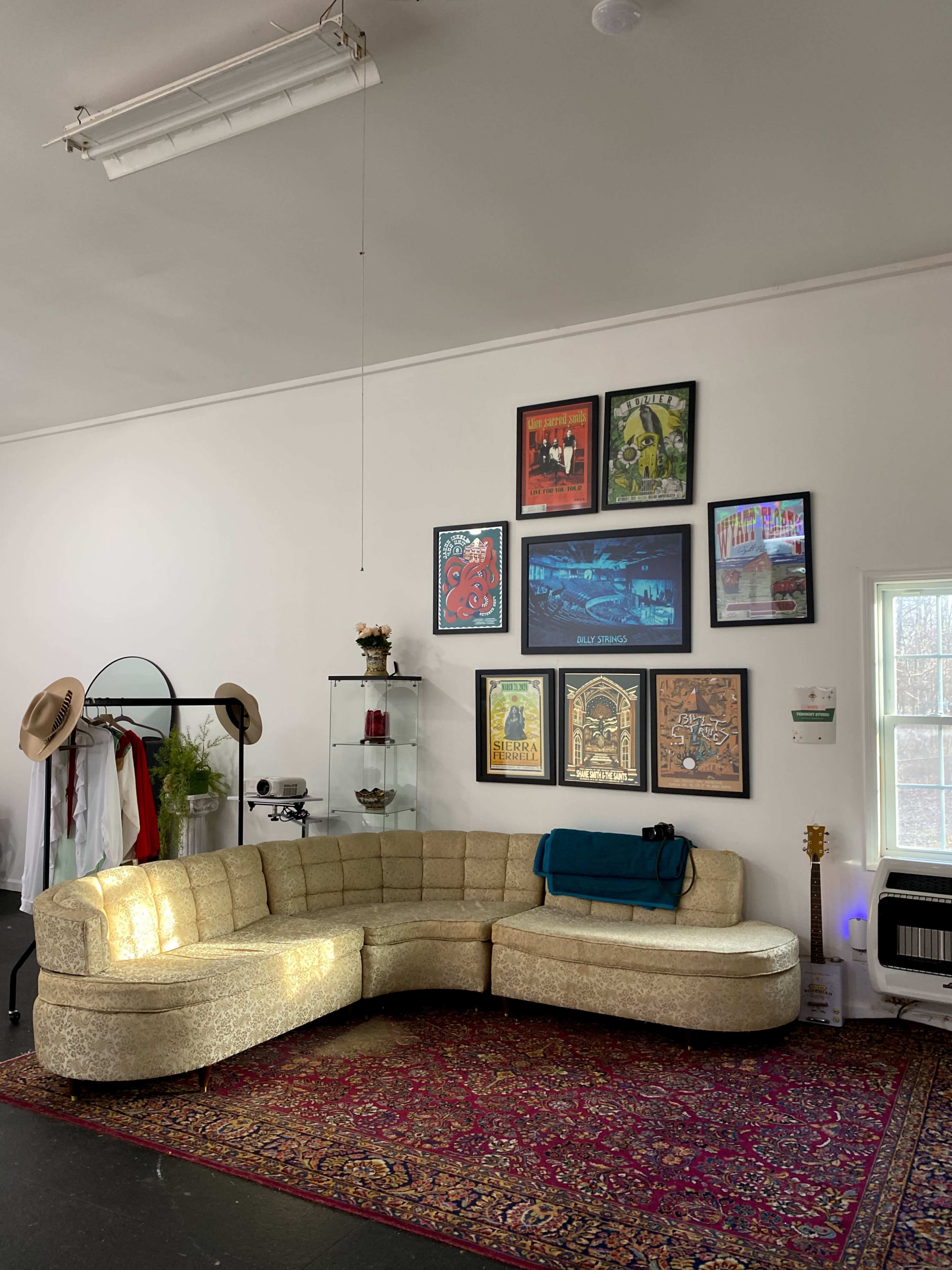 The image shows a spacious room featuring a gold sectional sofa against a white wall adorned with various framed posters, alongside a clothing rack and a guitar leaning against a wall.