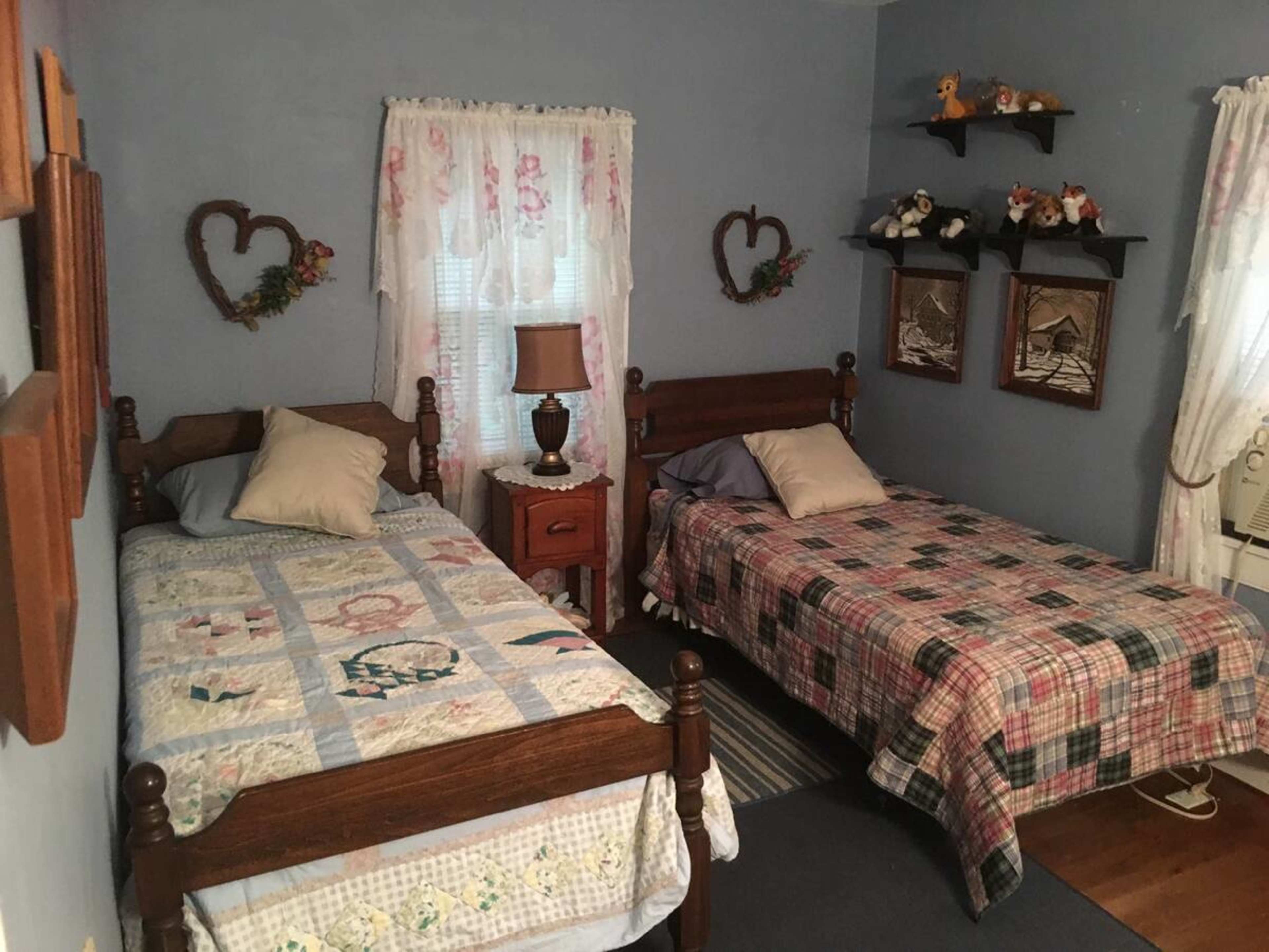 The image shows a small guest room featuring two wooden twin beds with colorful quilts, a bedside table with a lamp, and decorative wall accents.