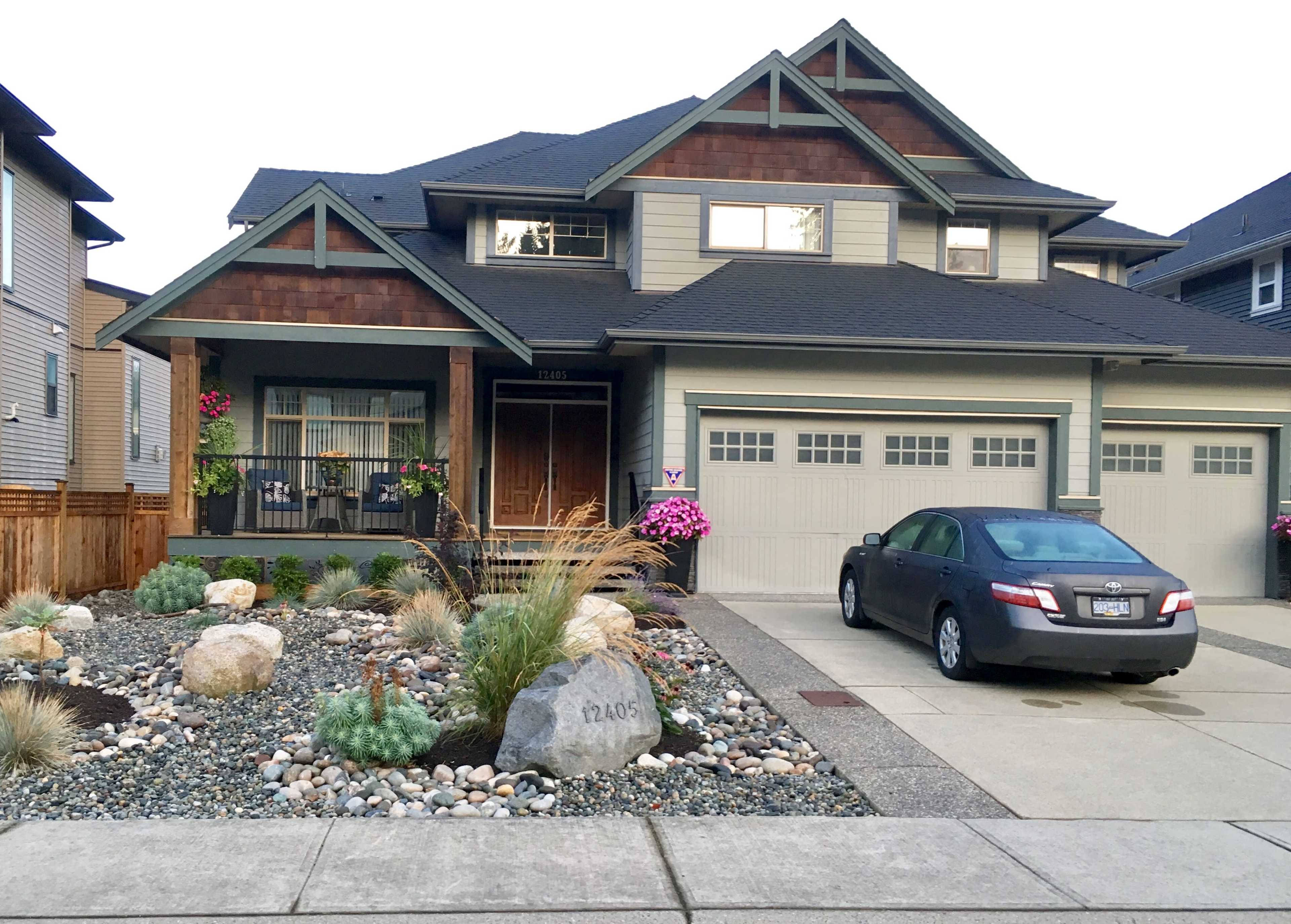 A two-story house with a mix of wood and siding features a landscaped front yard with rocks and plants, and a gray car parked in the driveway.