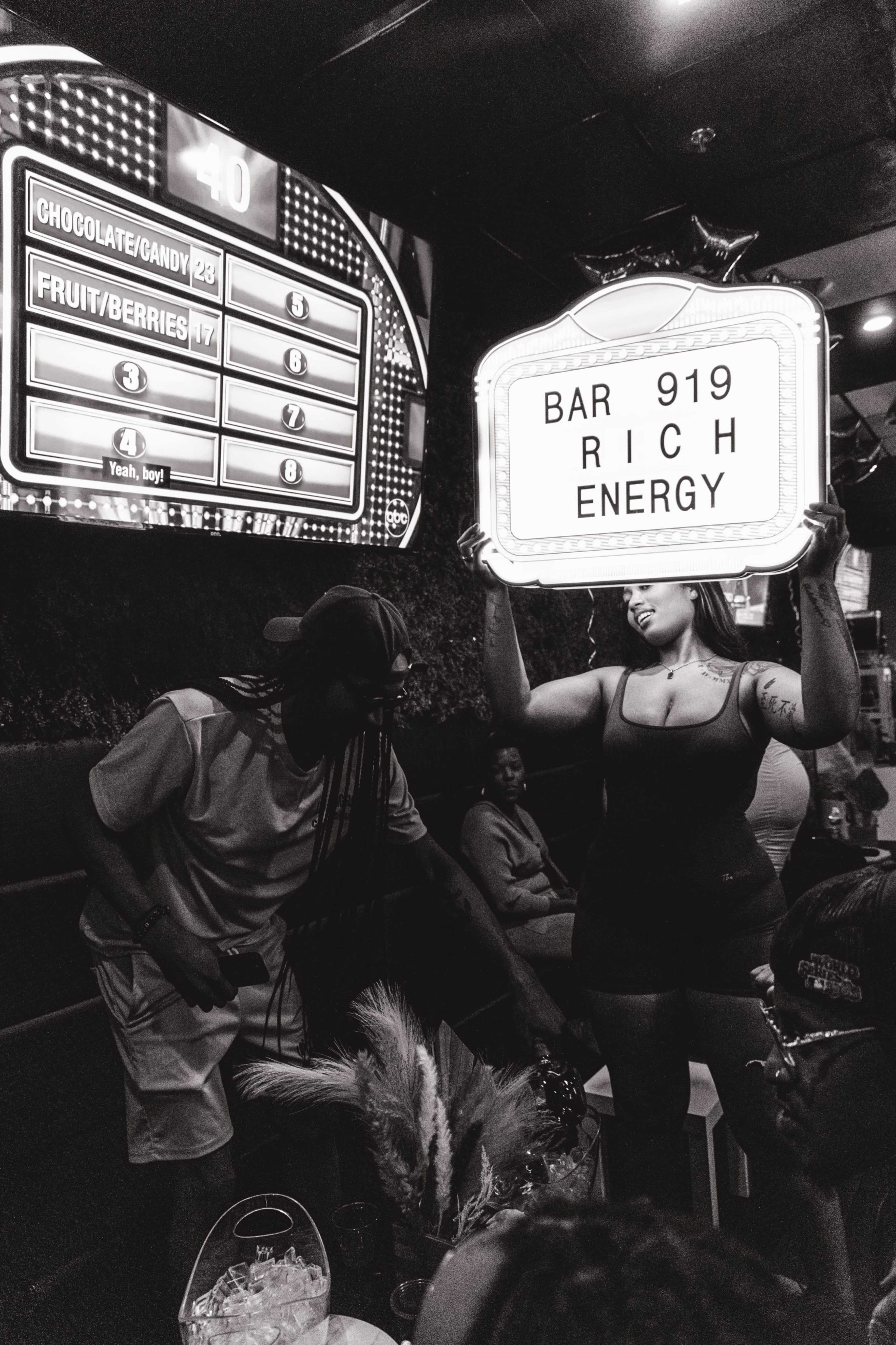A woman holds a sign reading "BAR 919 RICH ENERGY" while a group of people sits at a table in a dimly lit venue.