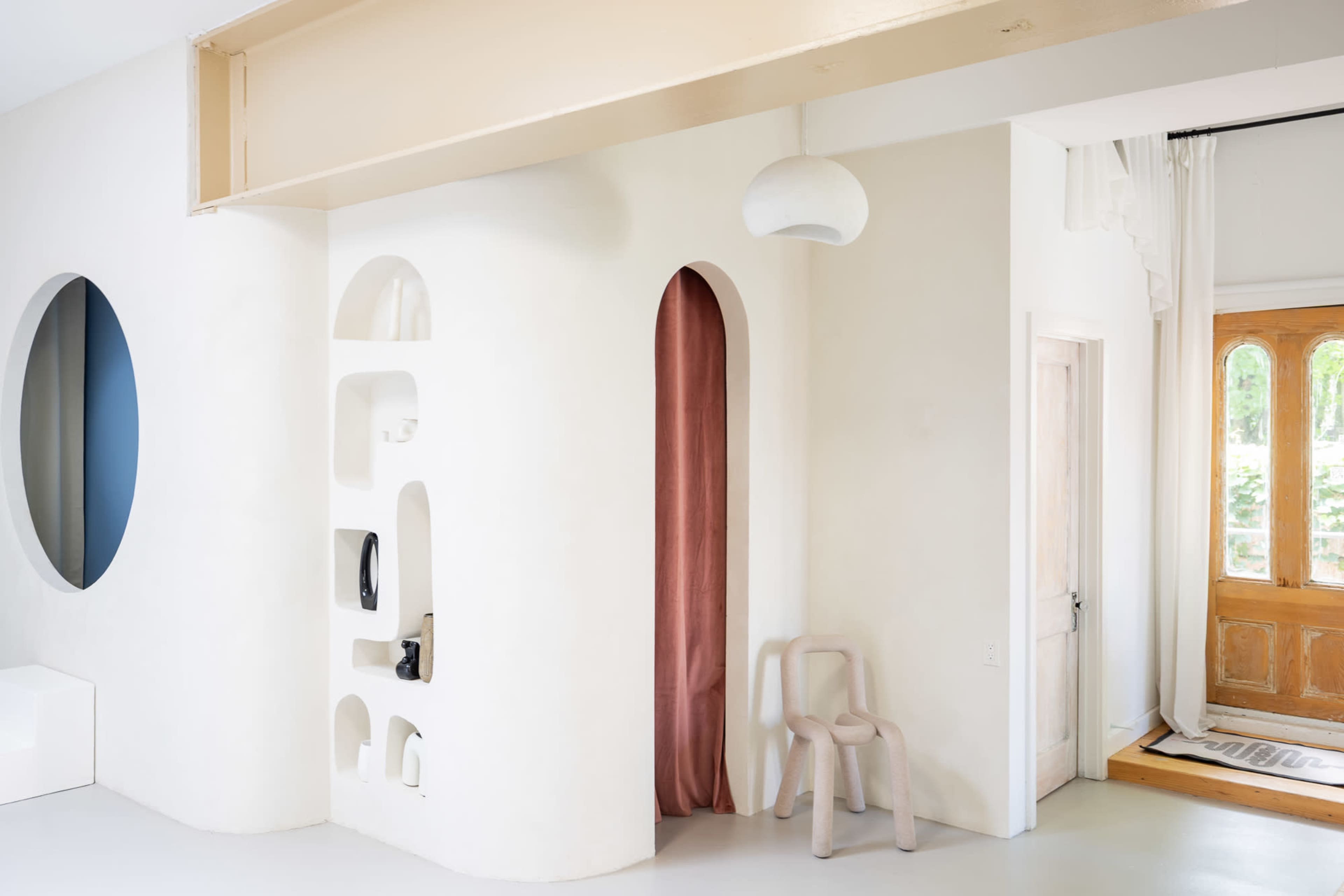 The image shows a modern interior space with a white wall featuring various alcoves, a partially concealed entryway draped with fabric, and a wooden door with glass panels.