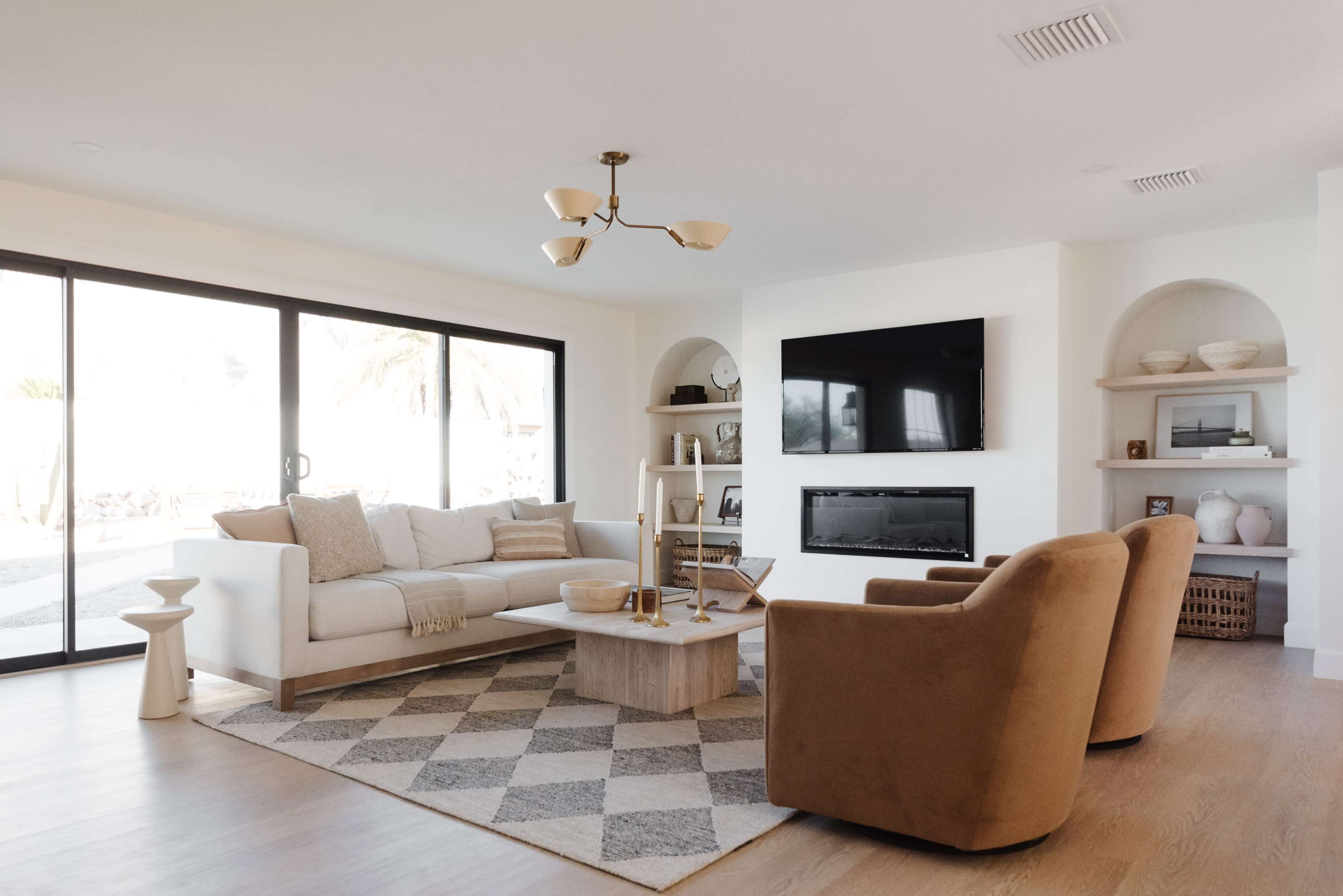 A modern living room features a beige couch and two brown armchairs, a coffee table on a geometric rug, and large glass doors leading to an outdoor area.