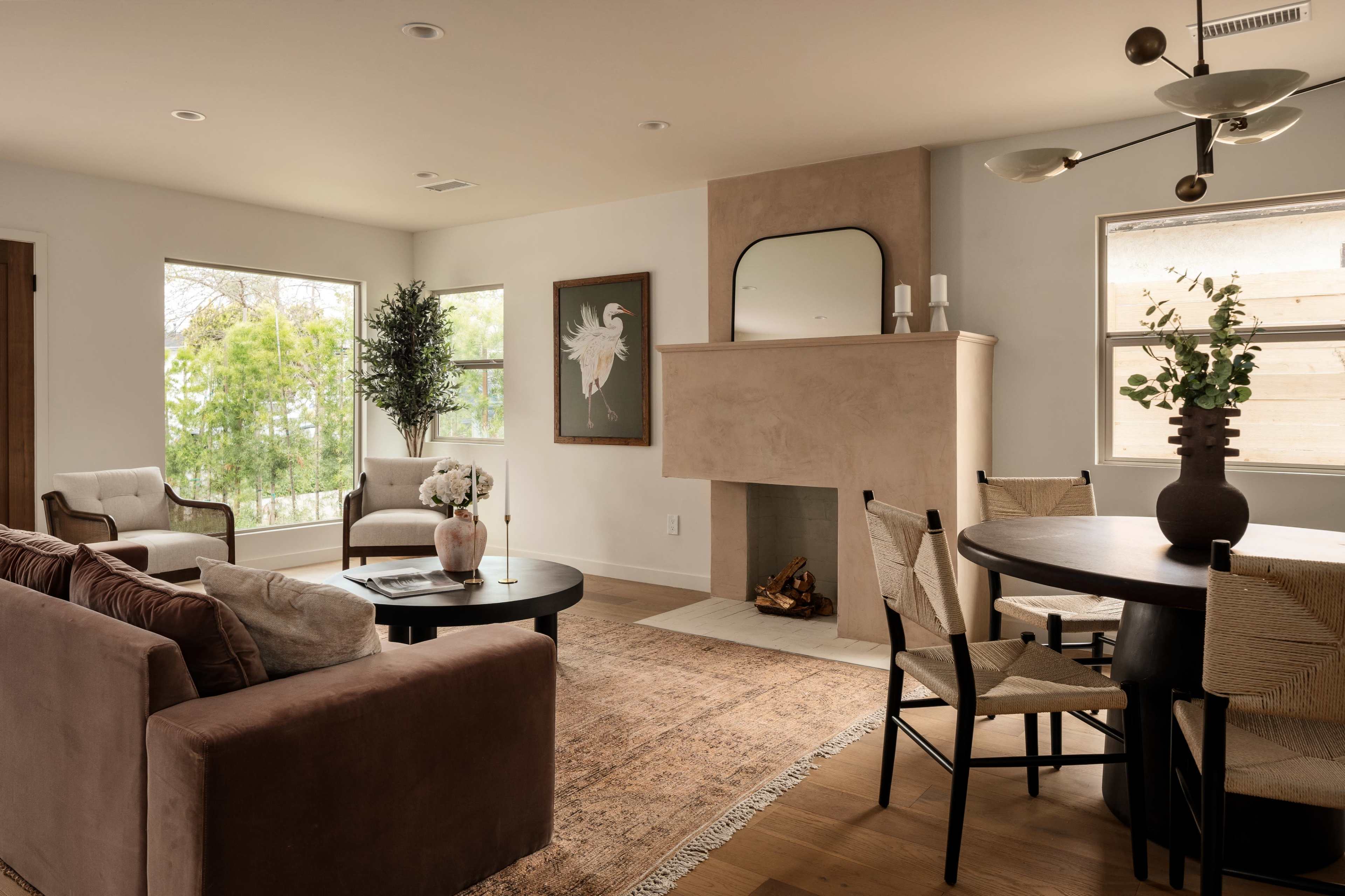 The living room features a large window, a comfortable sofa, a round black table, and a minimalist fireplace with a mirror above it.