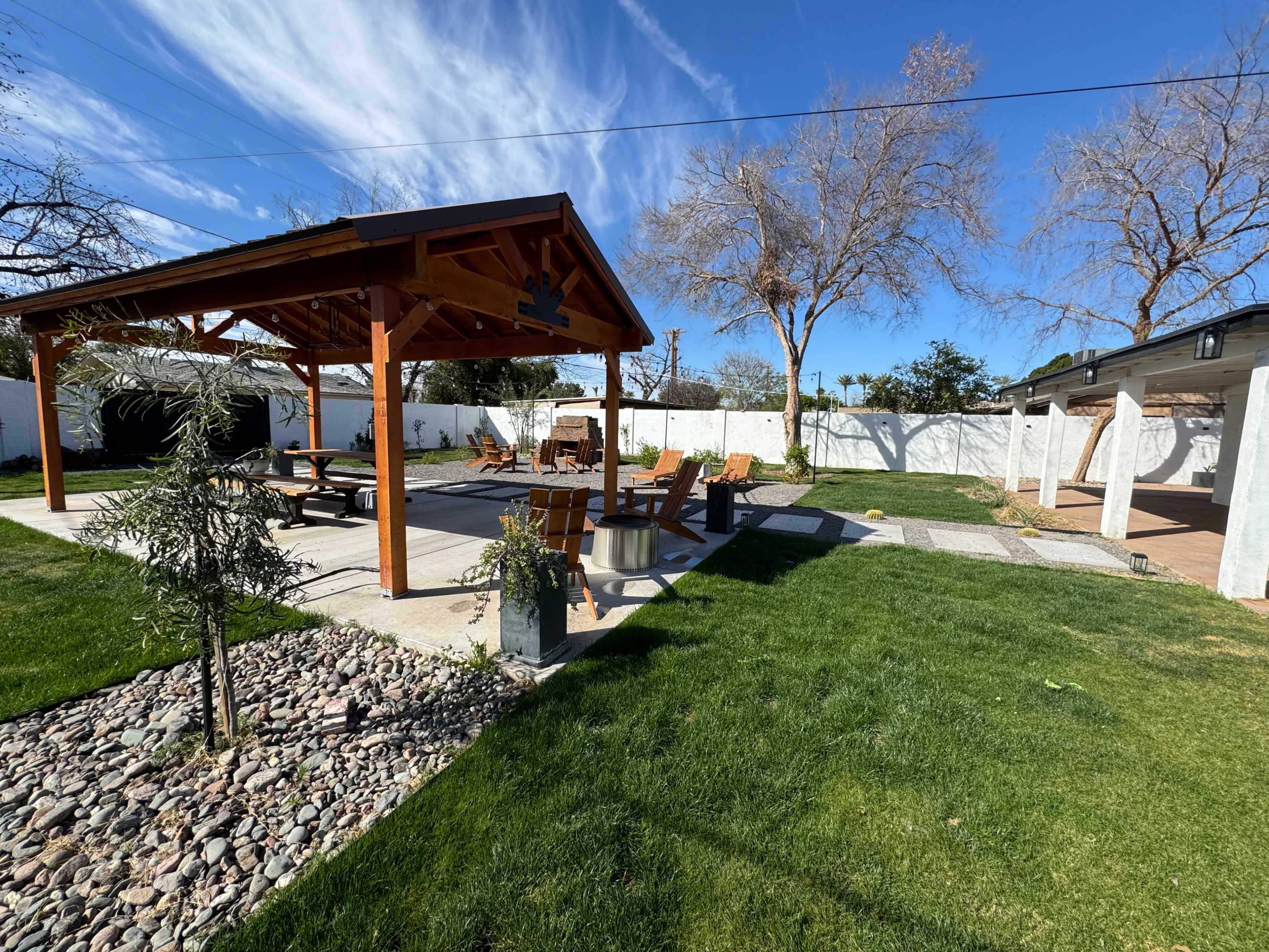 A backyard with a covered patio, several wooden chairs, a stone pathway, and landscaped grass areas.