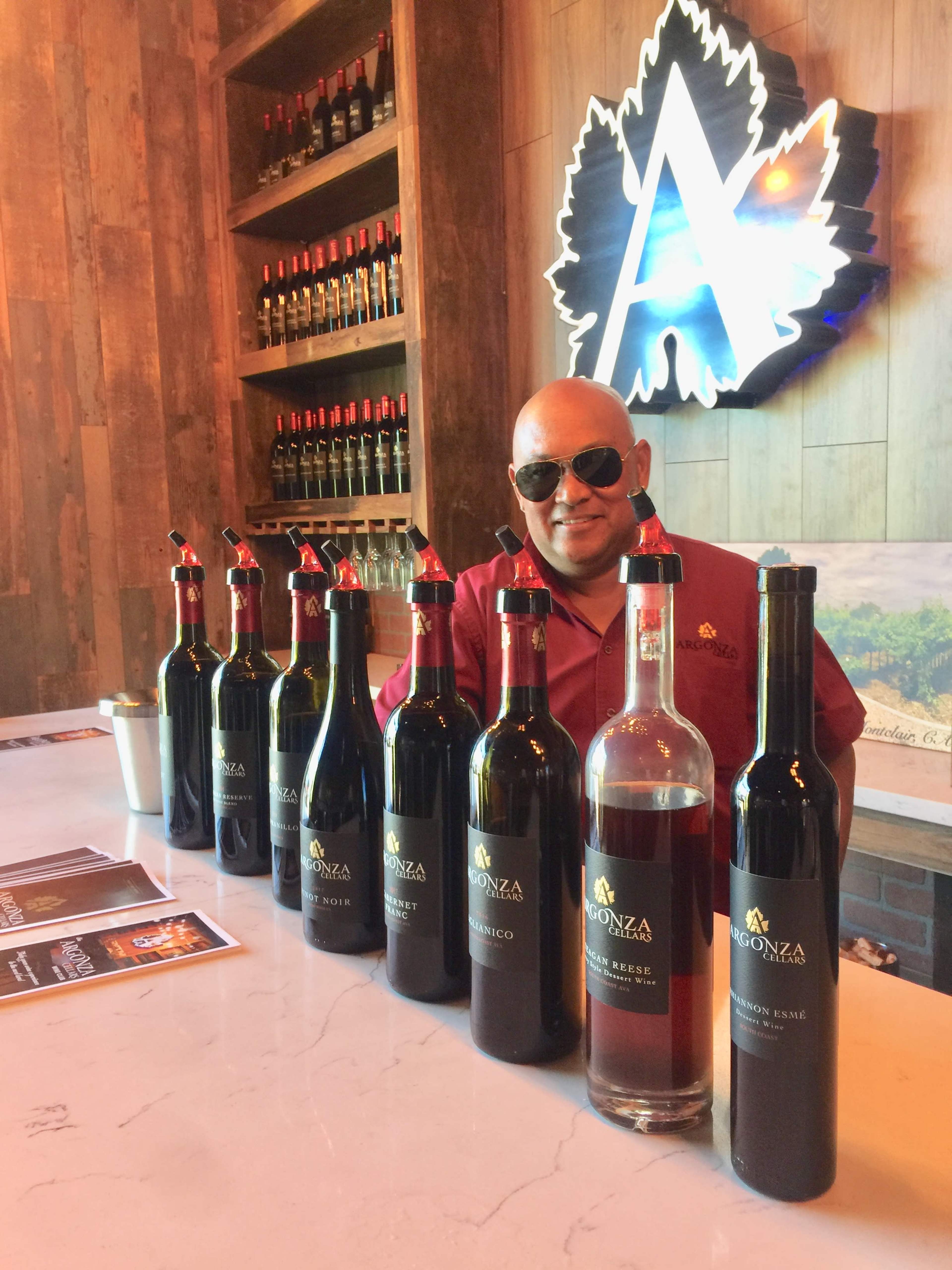 A man wearing sunglasses stands behind a bar showcasing several bottles of wine.