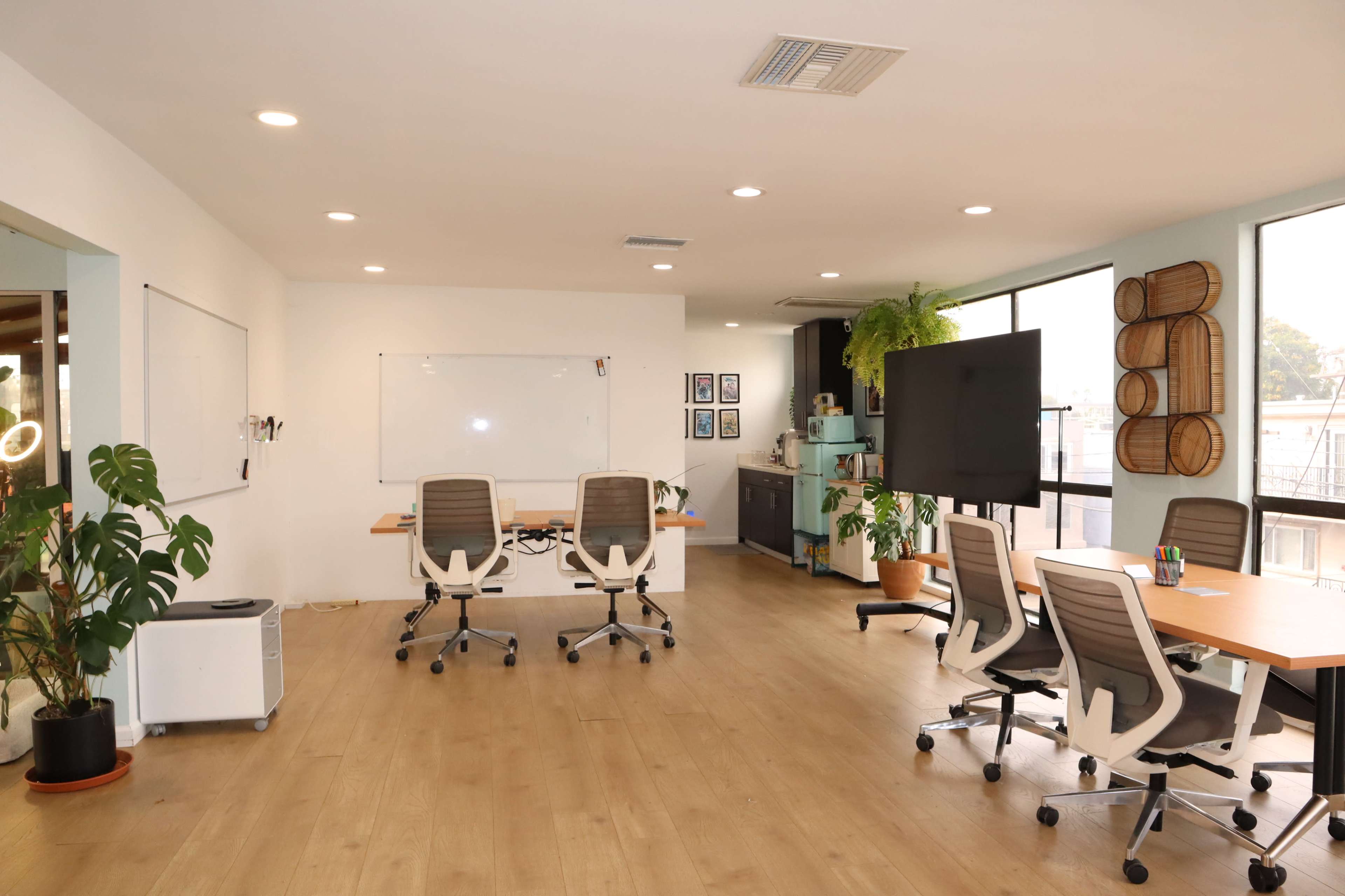 The image shows a modern office space featuring a whiteboard, two large work tables with chairs, and a television screen, all surrounded by natural light from large windows.
