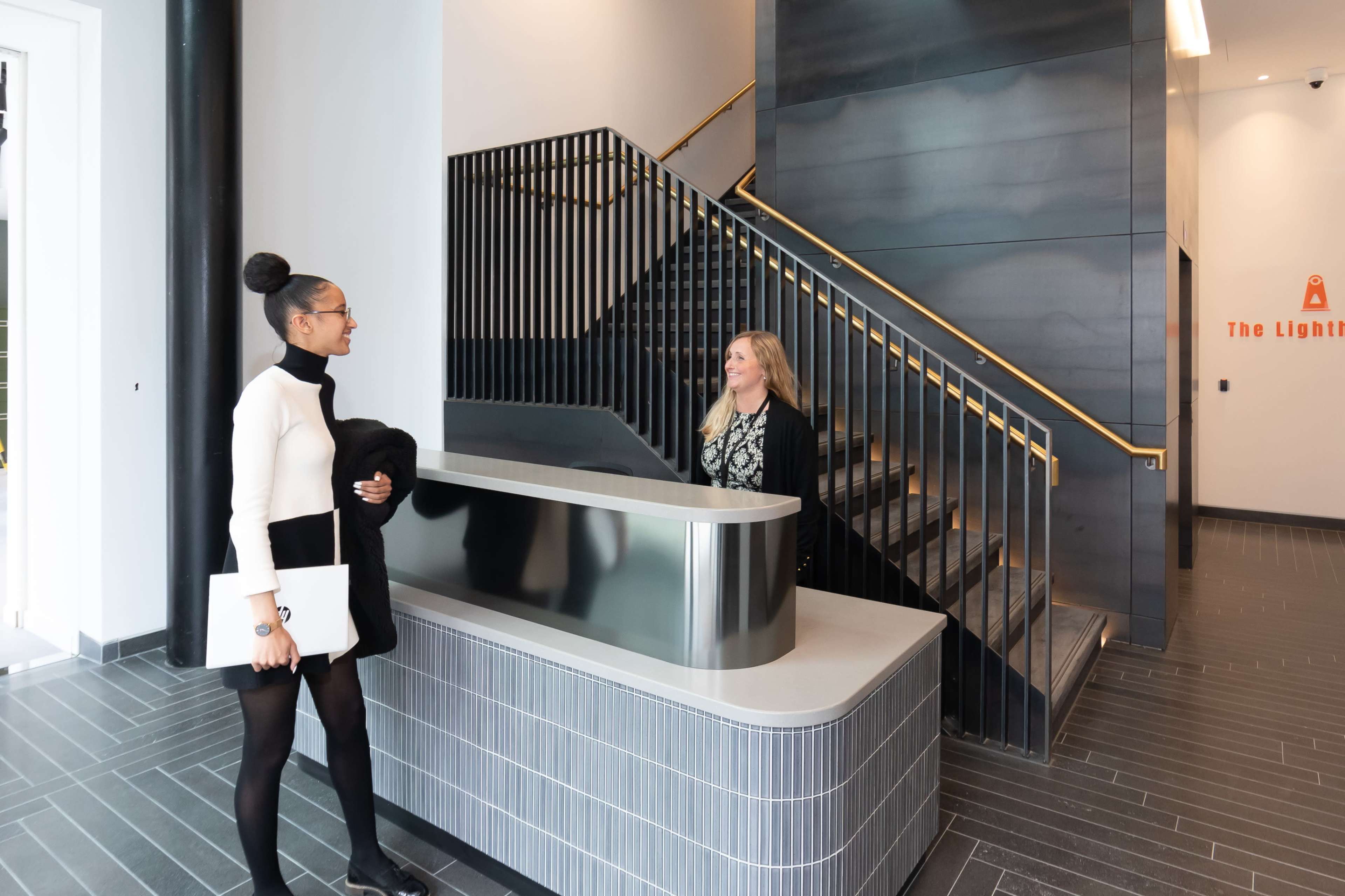 A woman in a black and white outfit stands at a reception desk while speaking to another woman near a staircase in a modern lobby.