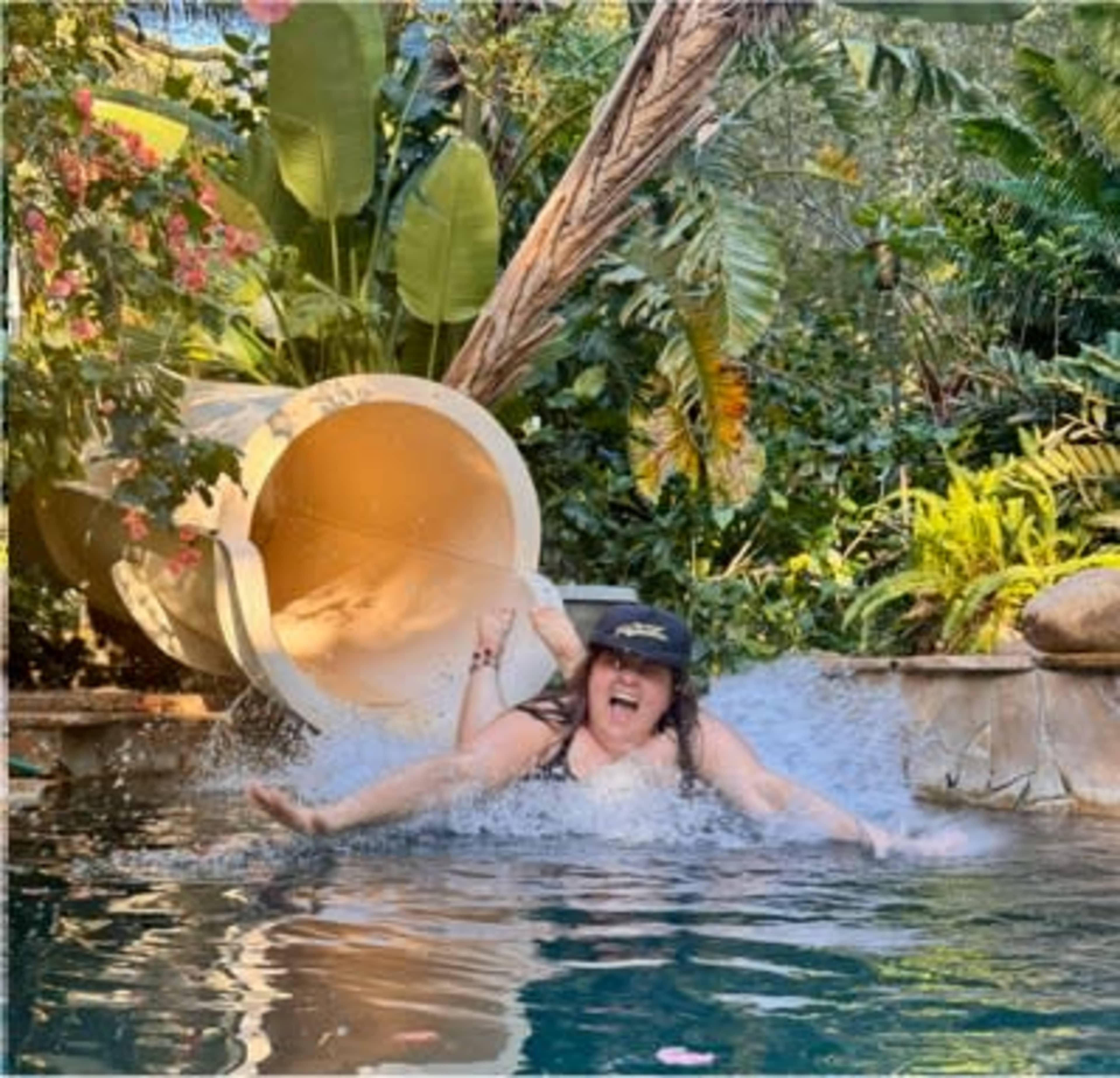 A person slides down a water slide into a swimming pool surrounded by tropical plants.