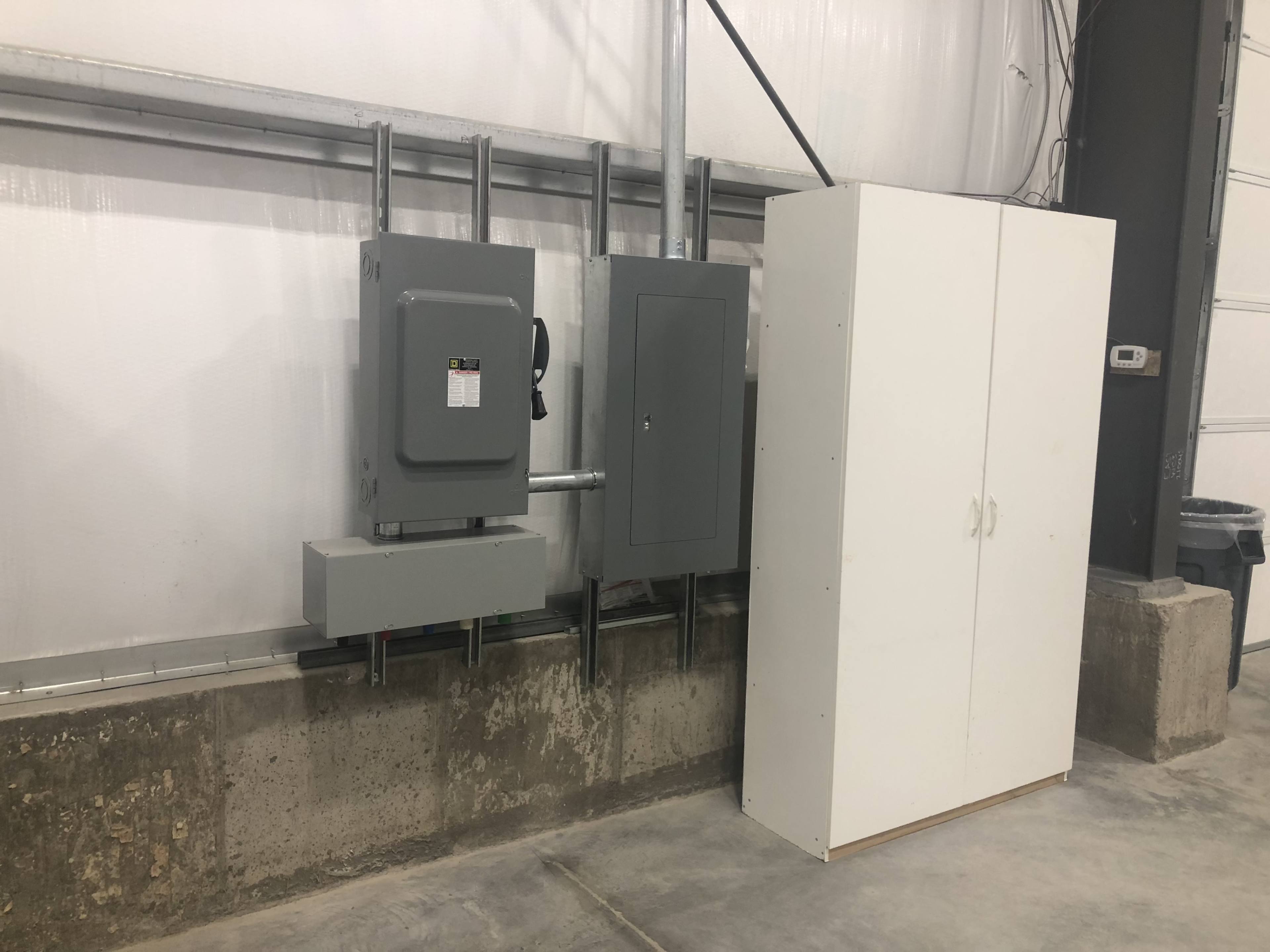 A gray electrical panel and a smaller gray box mounted on a concrete wall, next to a white storage cabinet in a facility.