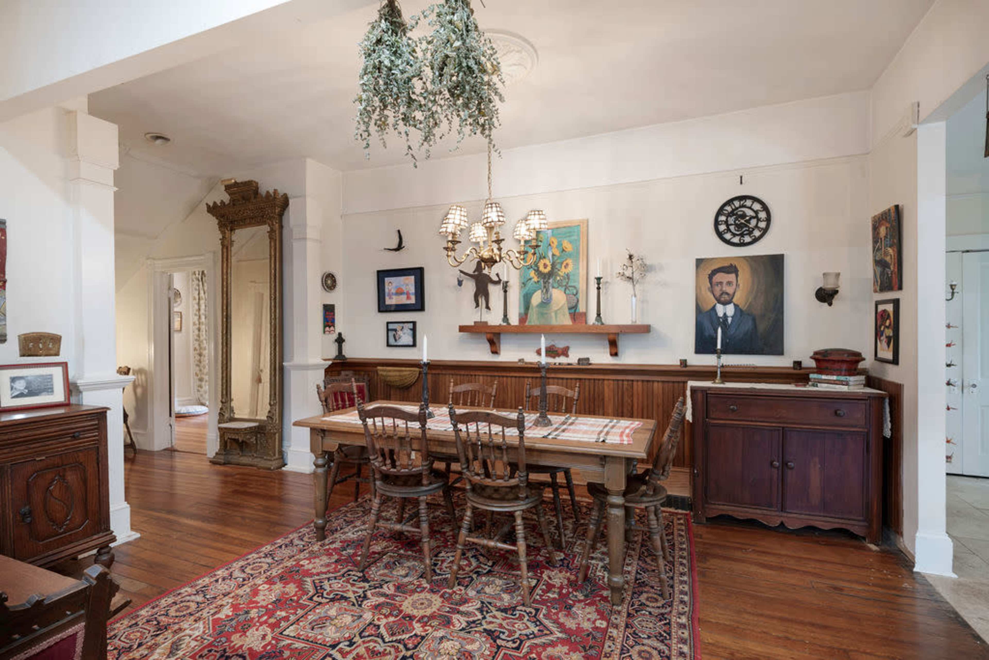 The image shows a dining area featuring a wooden table with chairs, a large mirror, and various artworks on the walls, all situated on hardwood flooring accented by a colorful rug.